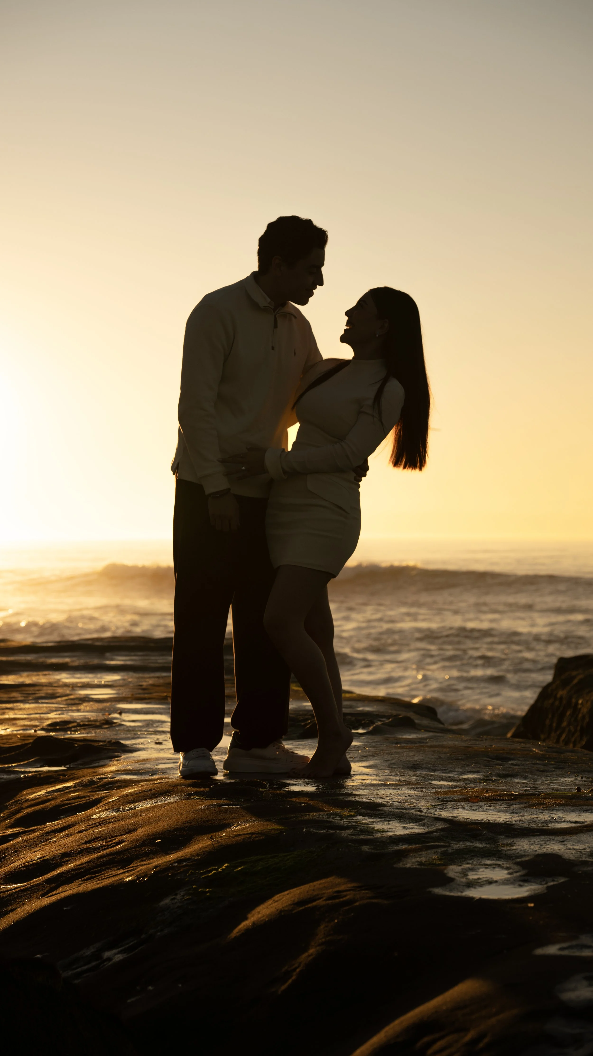 A silhouette of a man and woman standing close together on a beach at sunset, facing each other with the ocean in the background.