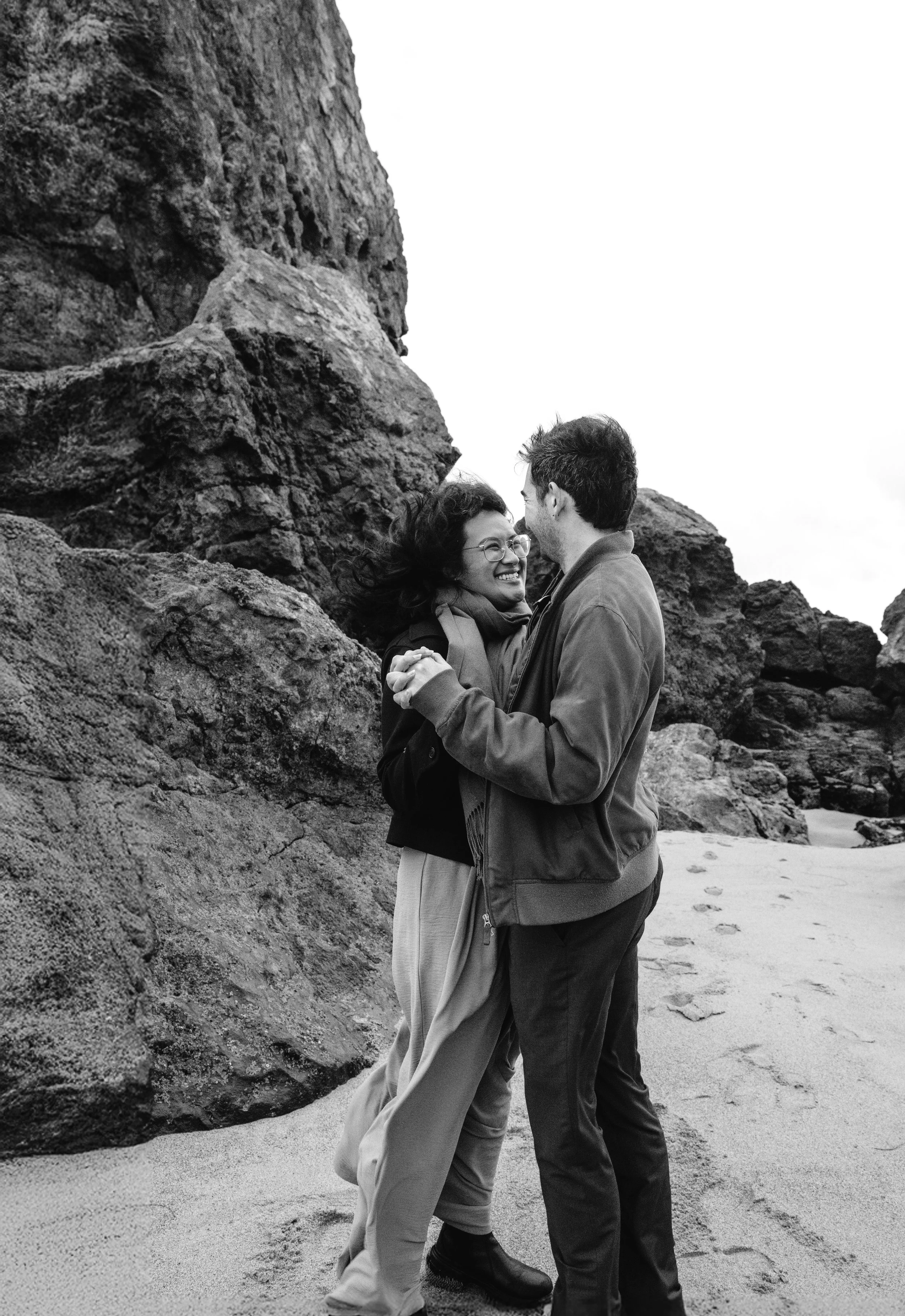 A couple dancing on a beach with large rocks in the background, smiling and holding hands.