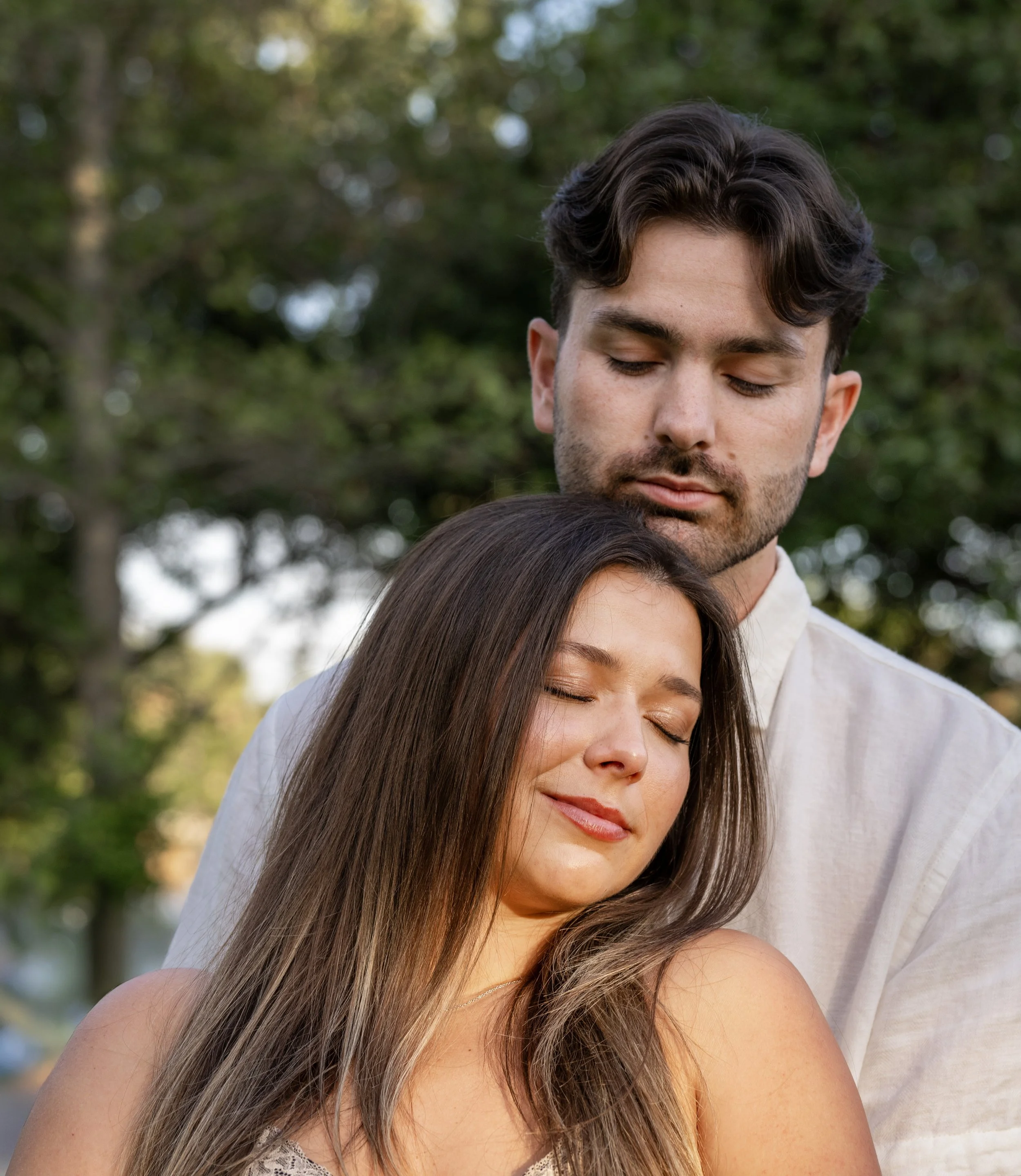 A young woman with long brown hair and closed eyes leaning on a man's shoulder. The man has dark hair, a beard, and is also with closed eyes. They are outdoors with a background of green trees.