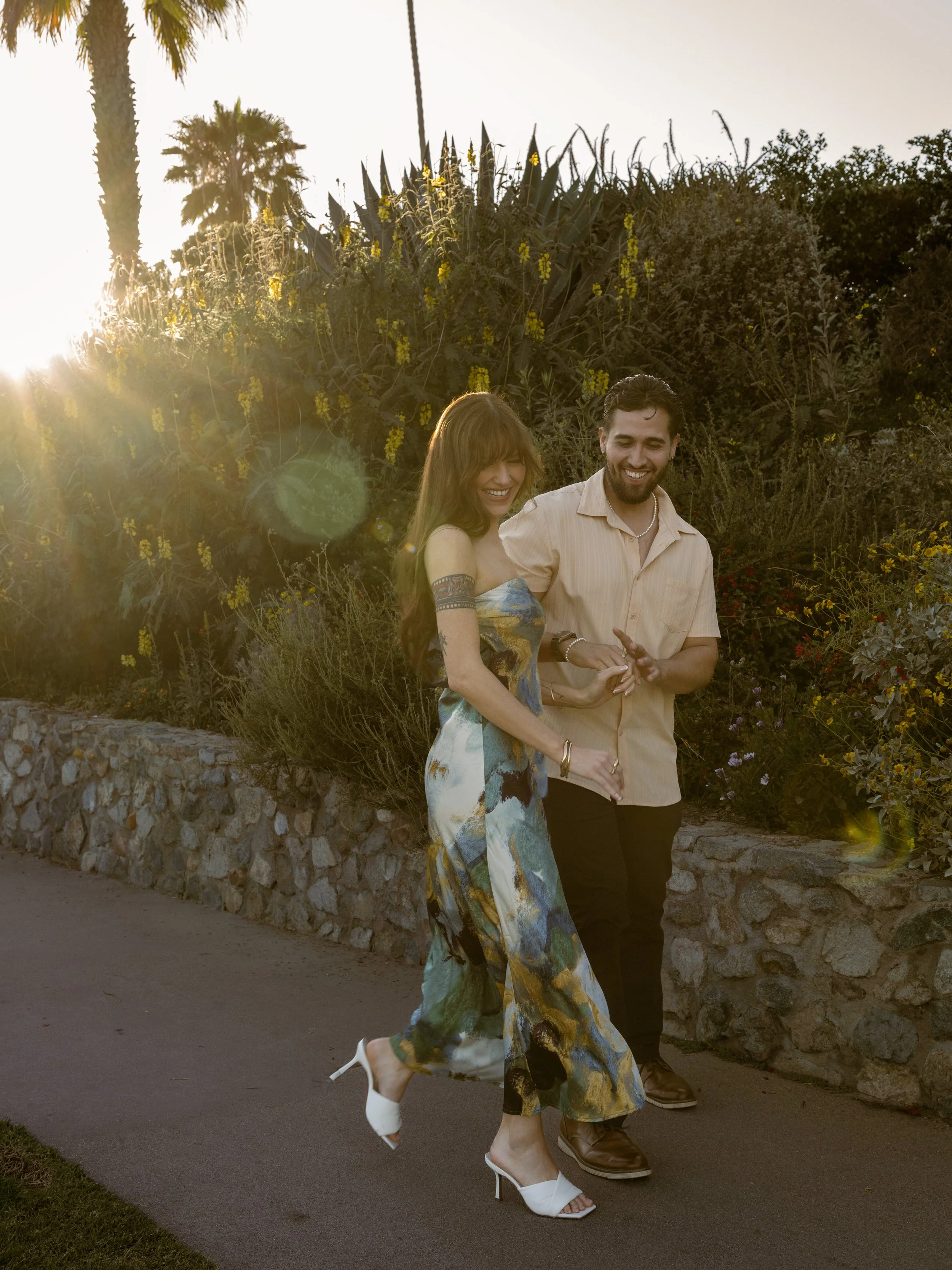 A couple walking outdoors at sunset, with the woman wearing a colorful dress and white heels and the man in a beige shirt and dark pants, smiling and holding hands.