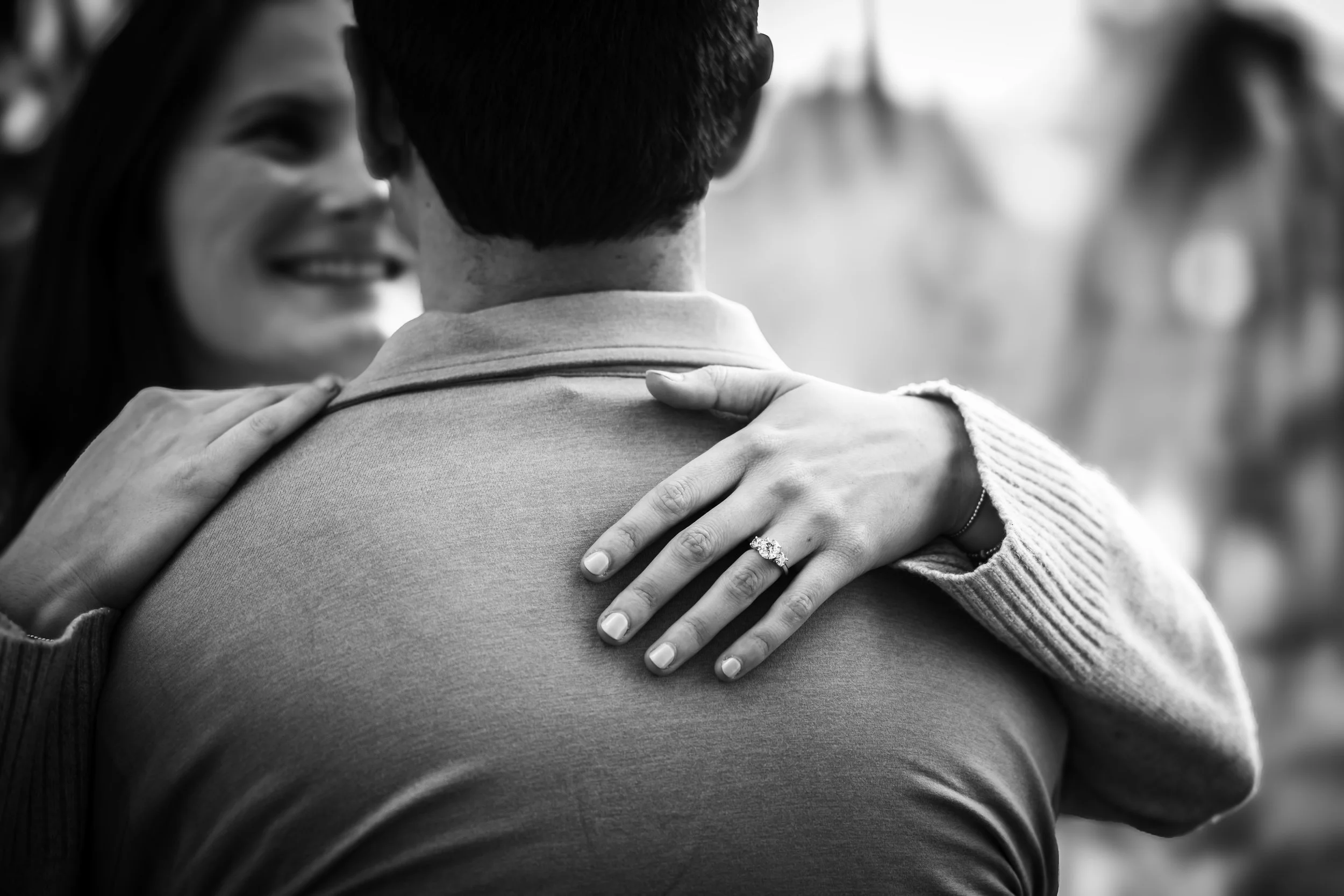 Black and white photo of a person with a woman embracing them from behind, her hands resting on their shoulders, with visible engagement ring on her finger, smiling and close to their neck, blurred background.