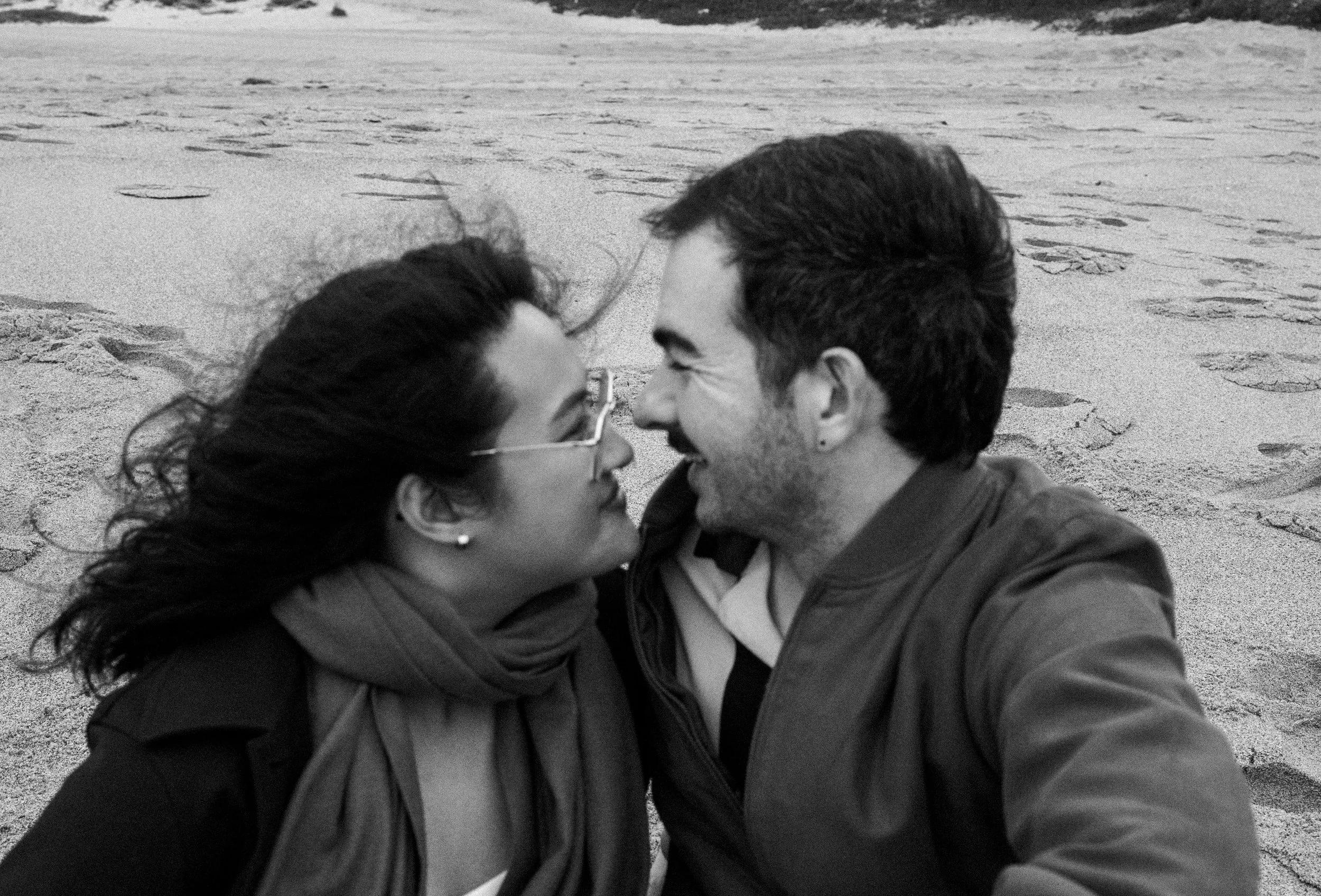 A black and white photo of a man and a woman close together on a beach, smiling and about to kiss.