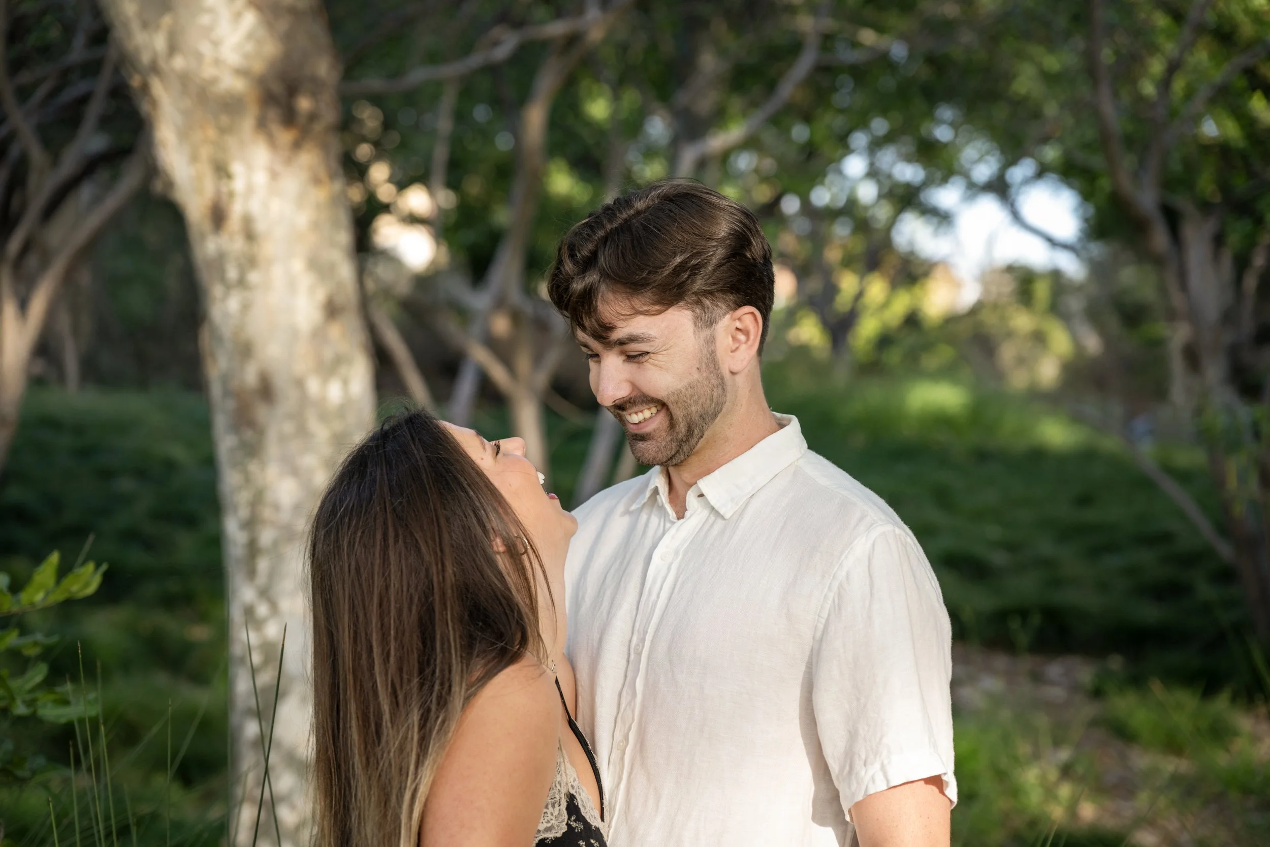 A smiling man and woman looking at each other outdoors in a green park with trees in the background.
