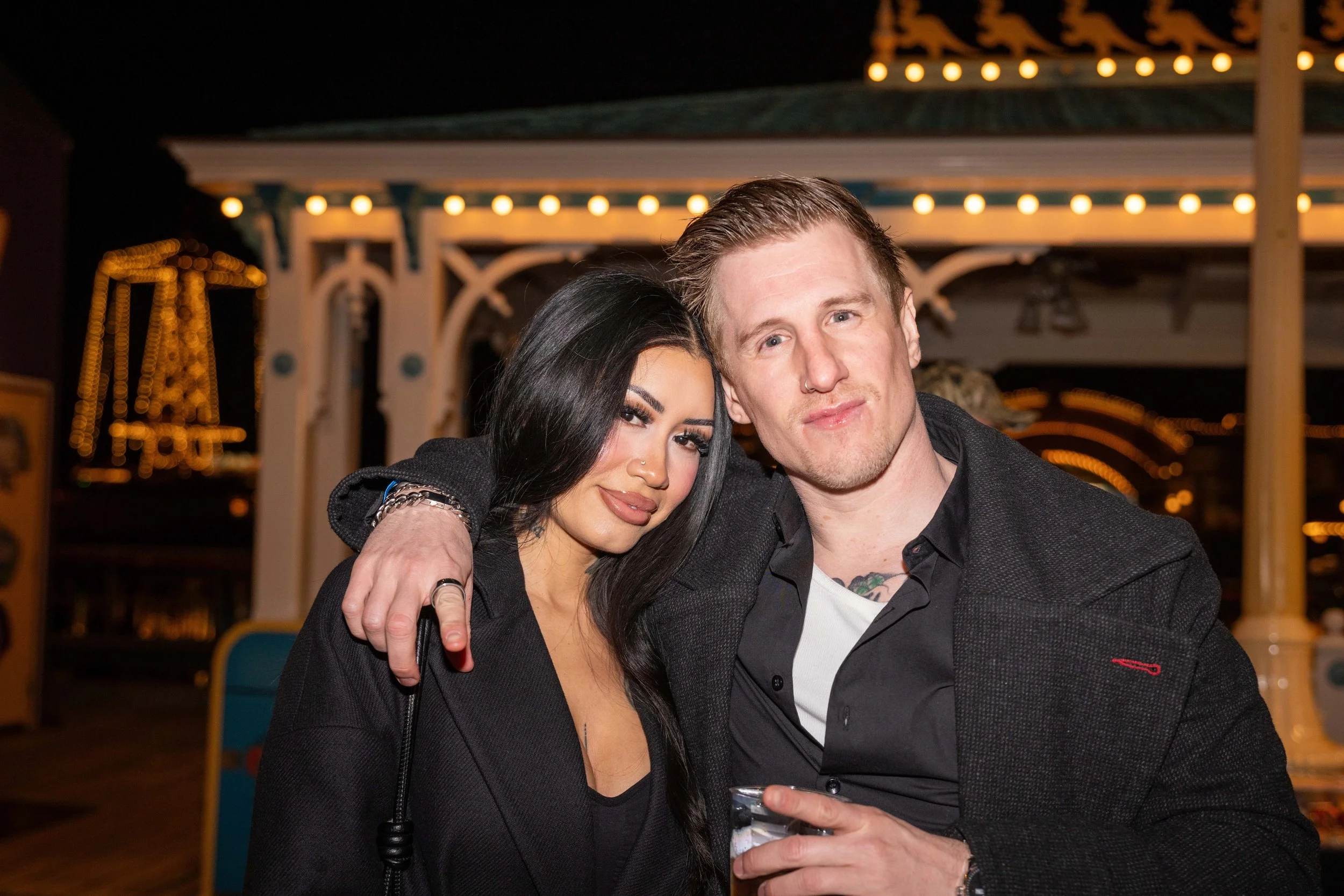 A man and woman posing closely together at night, with a decorative, lit-up background that appears to be an amusement park or carnival.