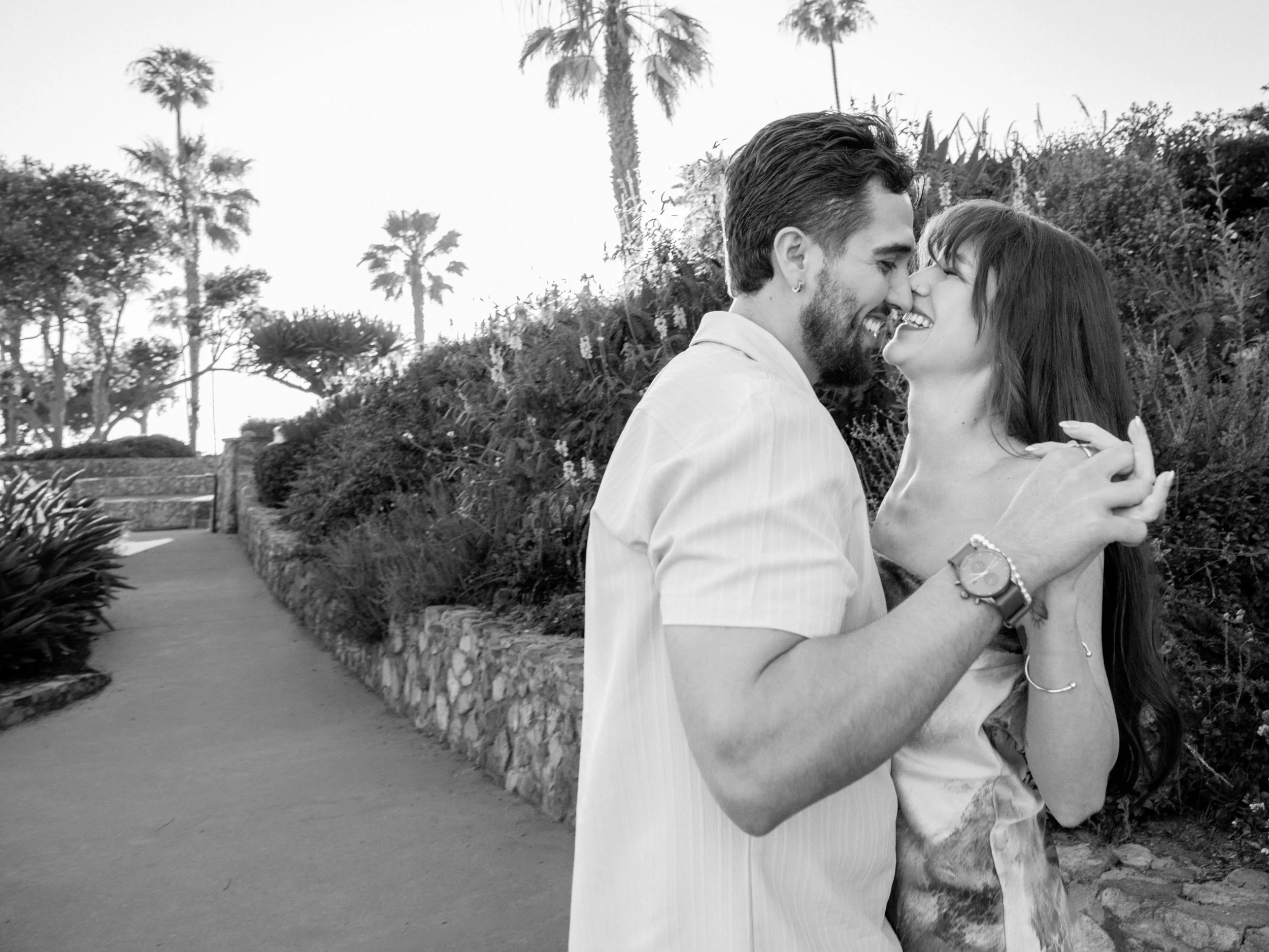 A black and white photo of a smiling couple in an outdoor garden setting, about to kiss.