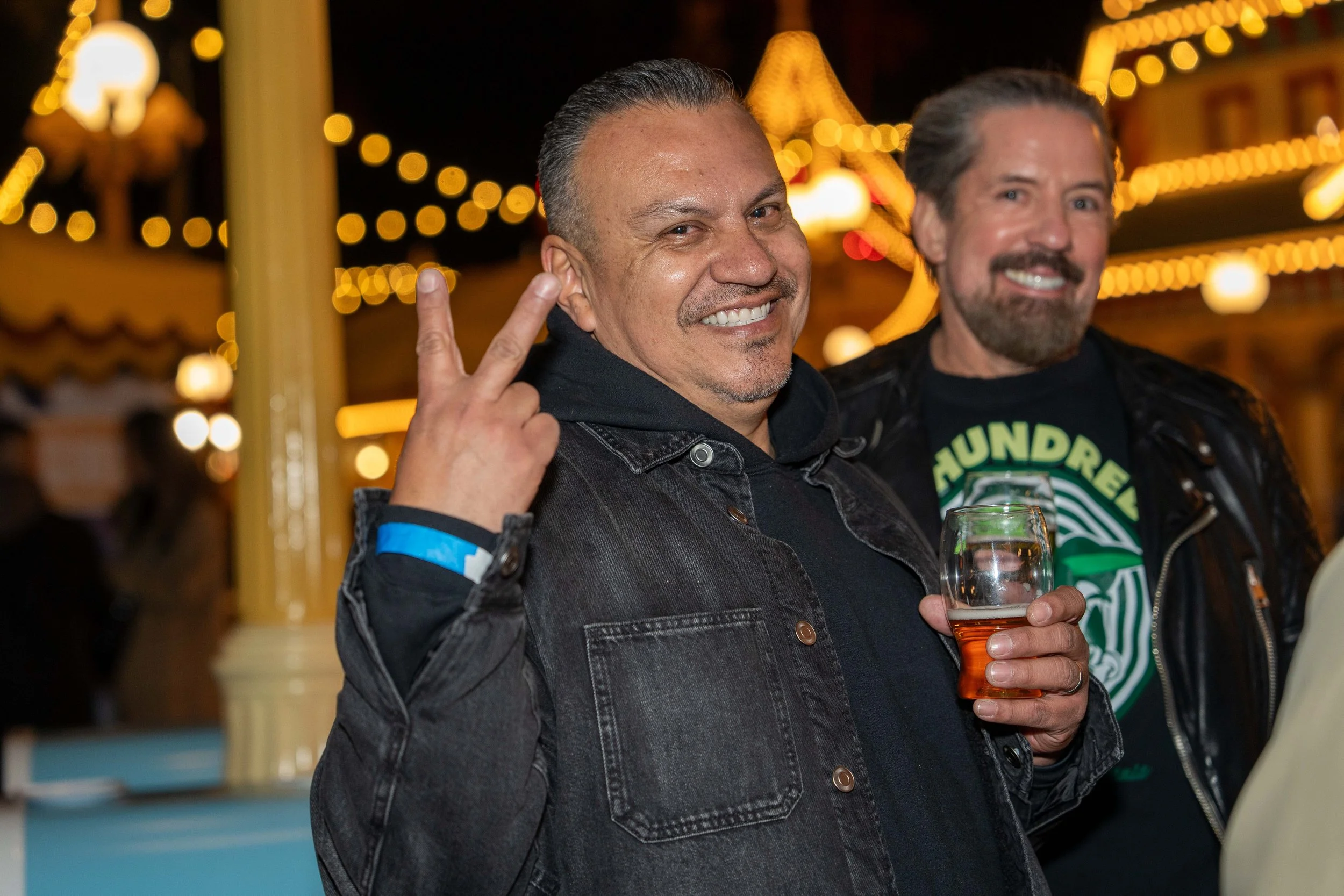 Two men smiling at a nighttime outdoor event, one making a peace sign and holding a glass of beer, with bright string lights in the background.
