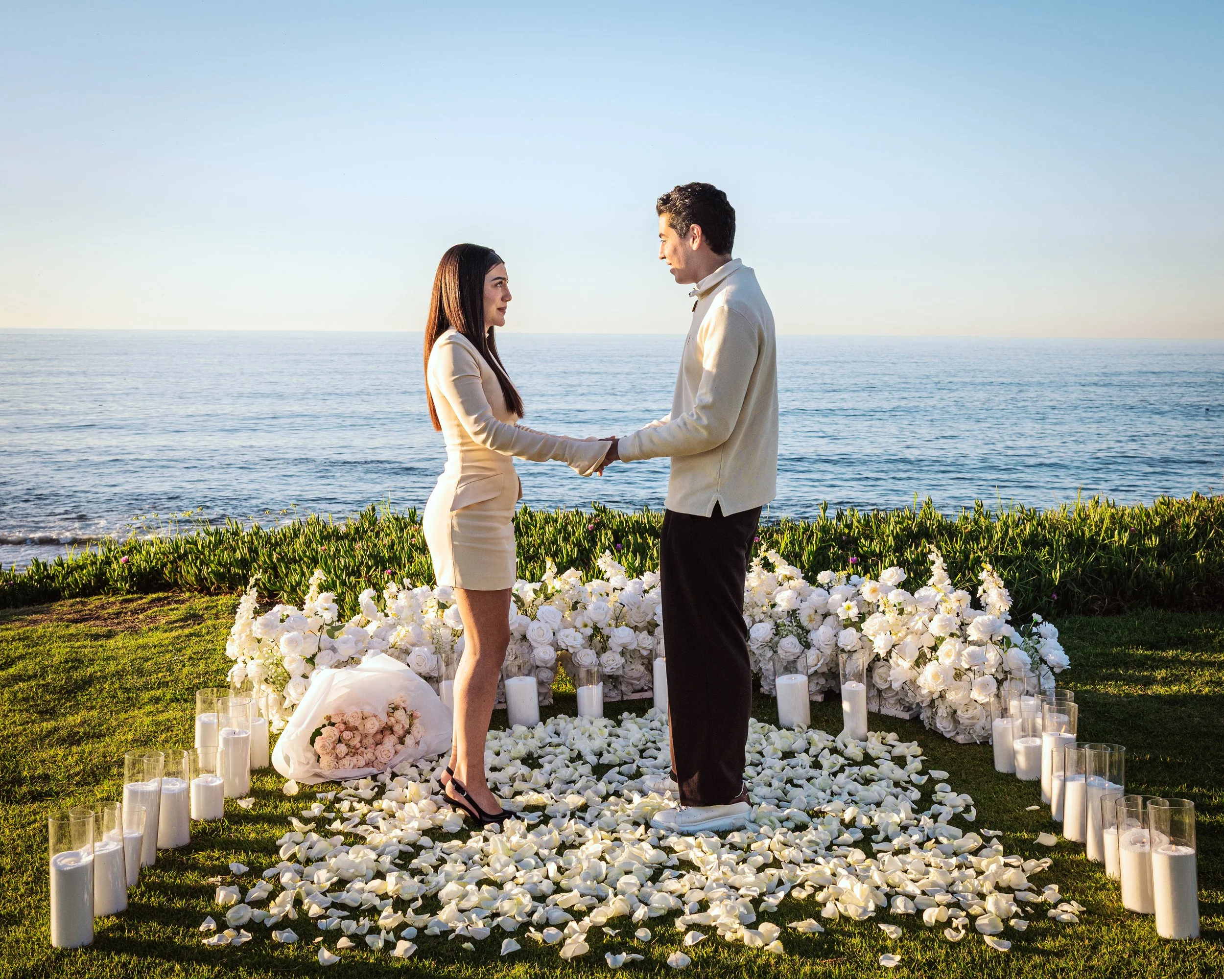A couple standing at an outdoor wedding ceremony by the ocean, holding hands and facing each other. The ceremony is decorated with white flowers, candles, and rose petals, with the sea in the background and the sky clear.