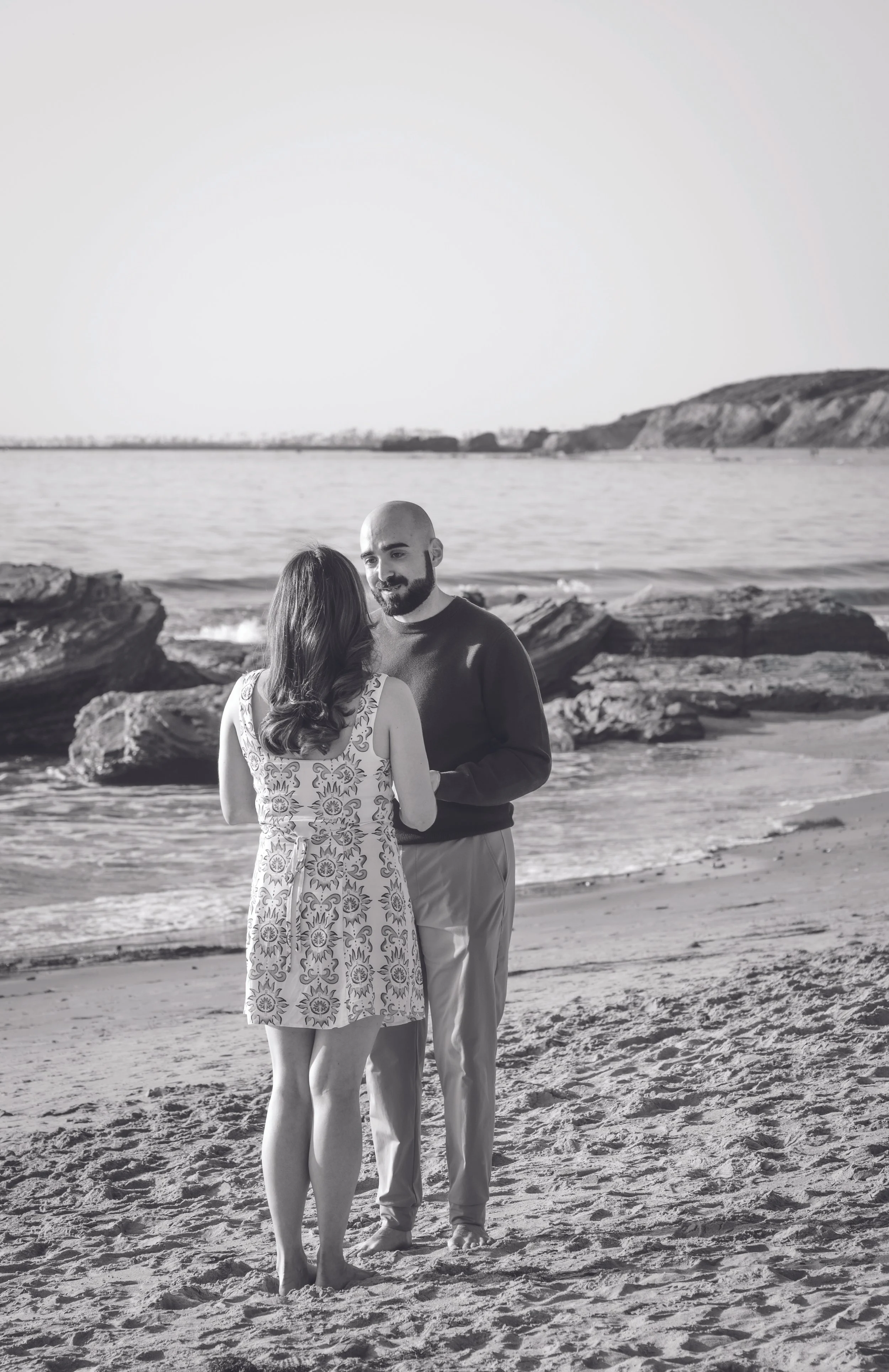 A man and woman standing on a sandy beach, facing each other, with ocean waves and rocky shoreline in the background.