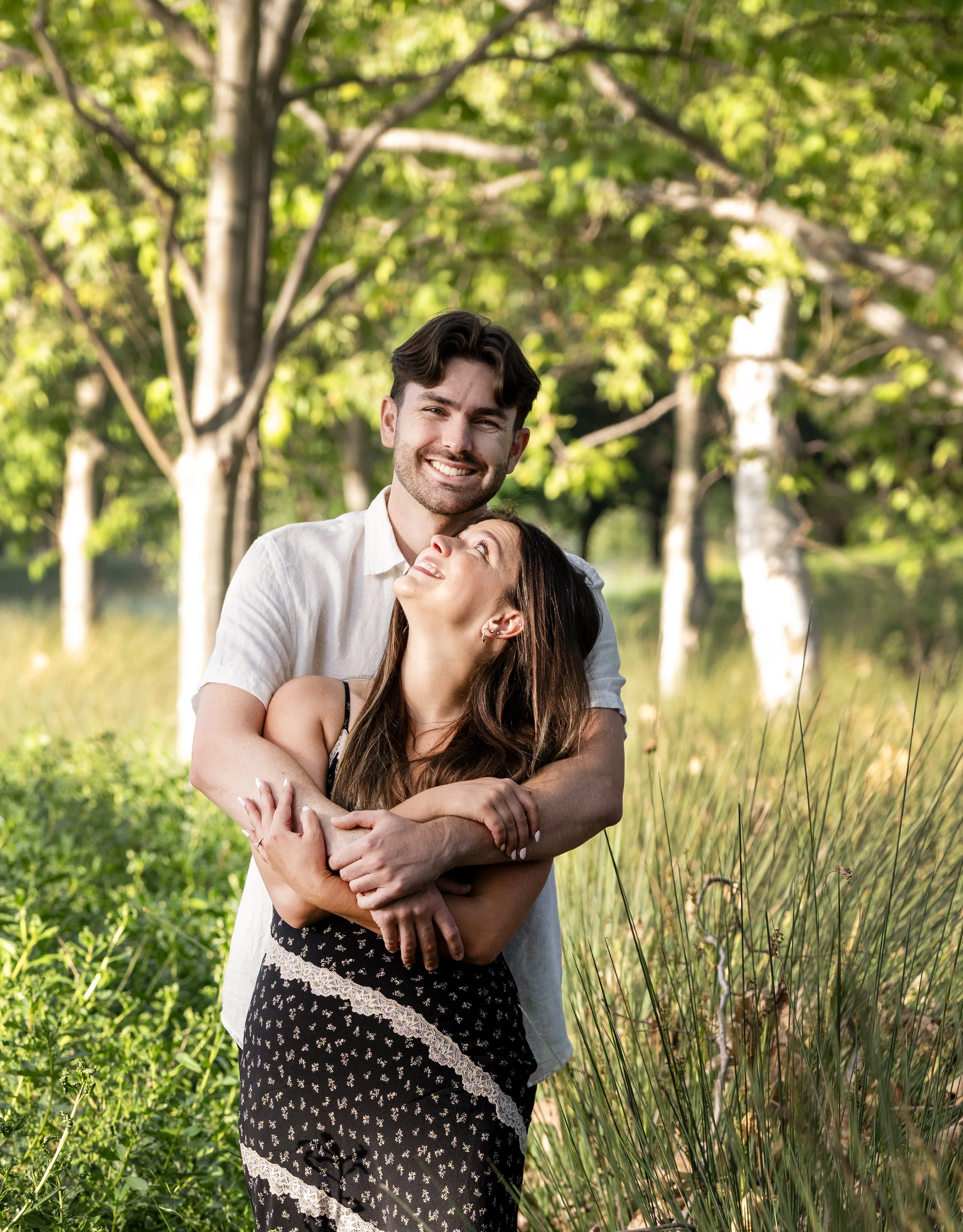 A happy couple embracing outdoors in a park with lush green trees and grass, sunny weather.