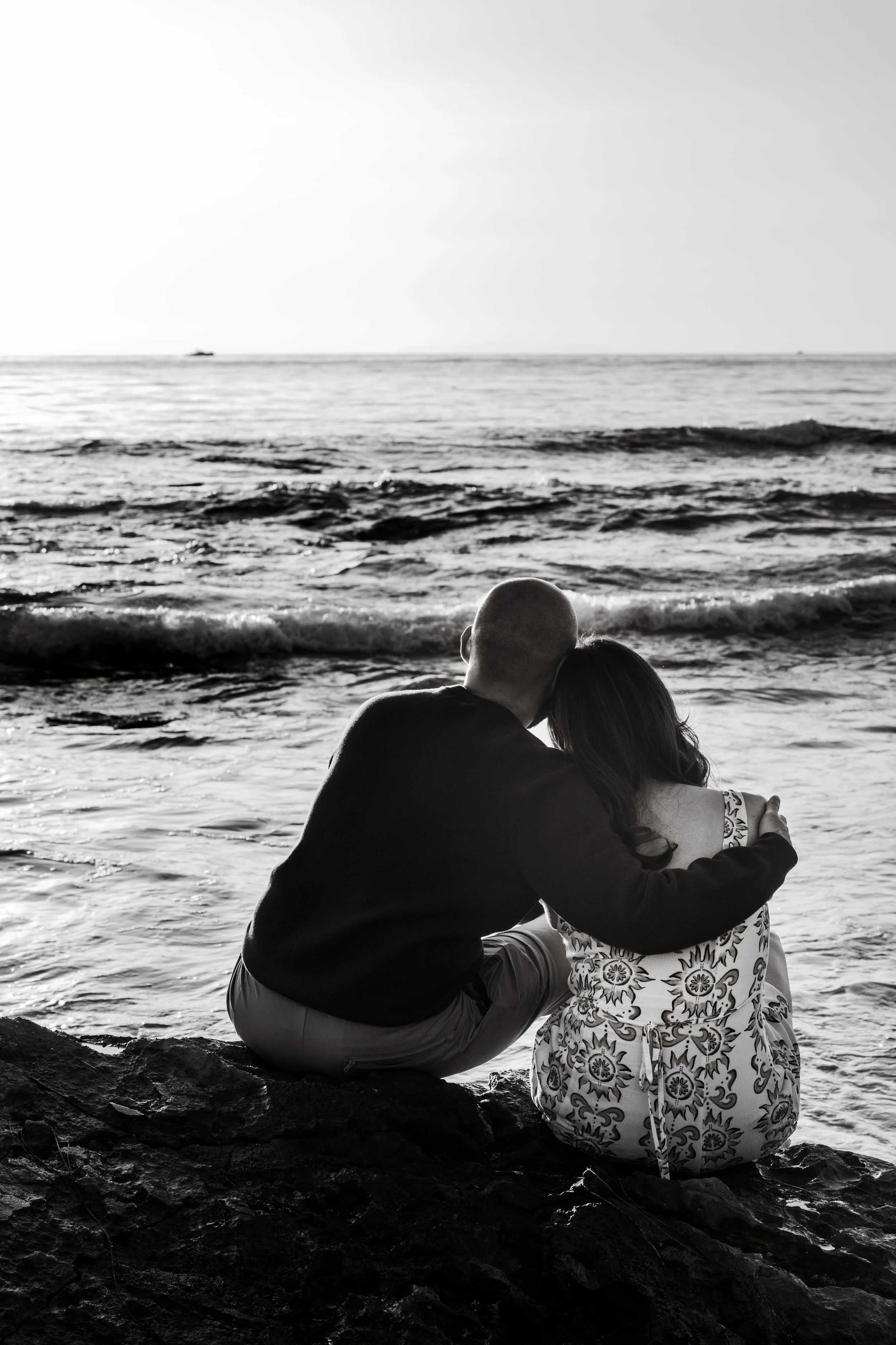 Black and white photo of a couple sitting on rocks at the beach, embracing and looking at the ocean, with a boat in the distance.