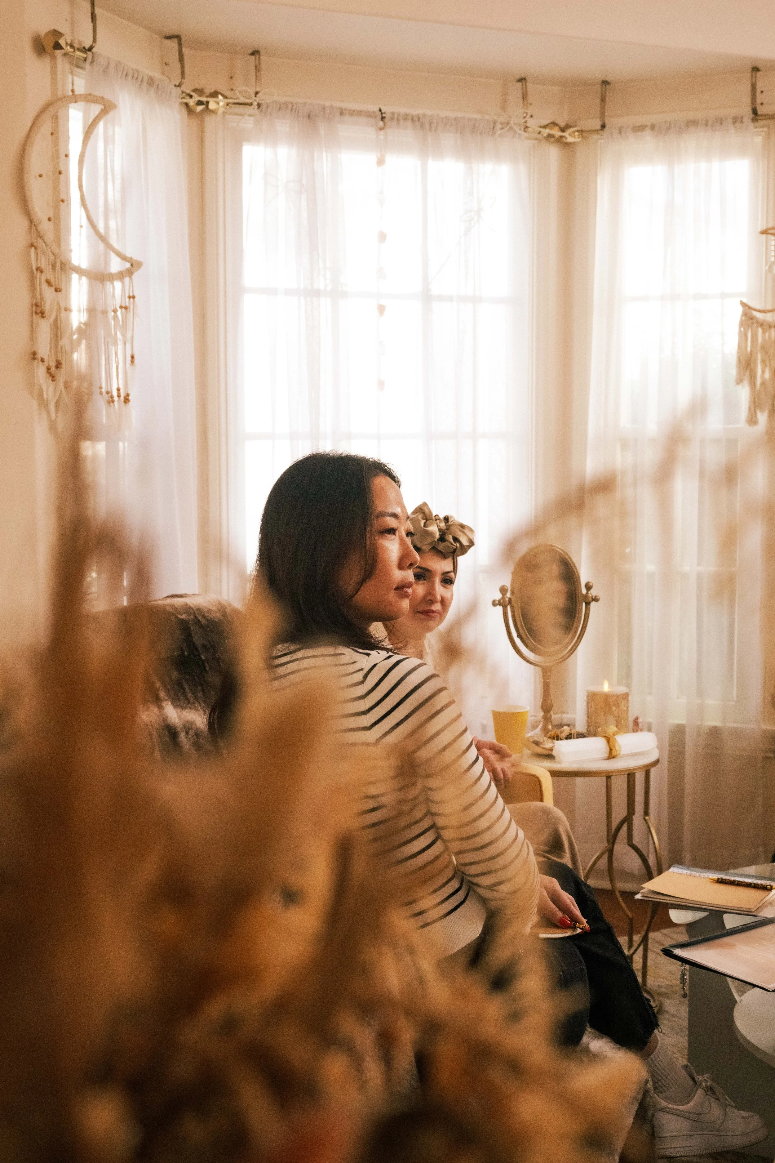 People sitting in a cozy room with natural light, engaged in conversation. The room is decorated with curtains, dream catchers, and candles on a side table.