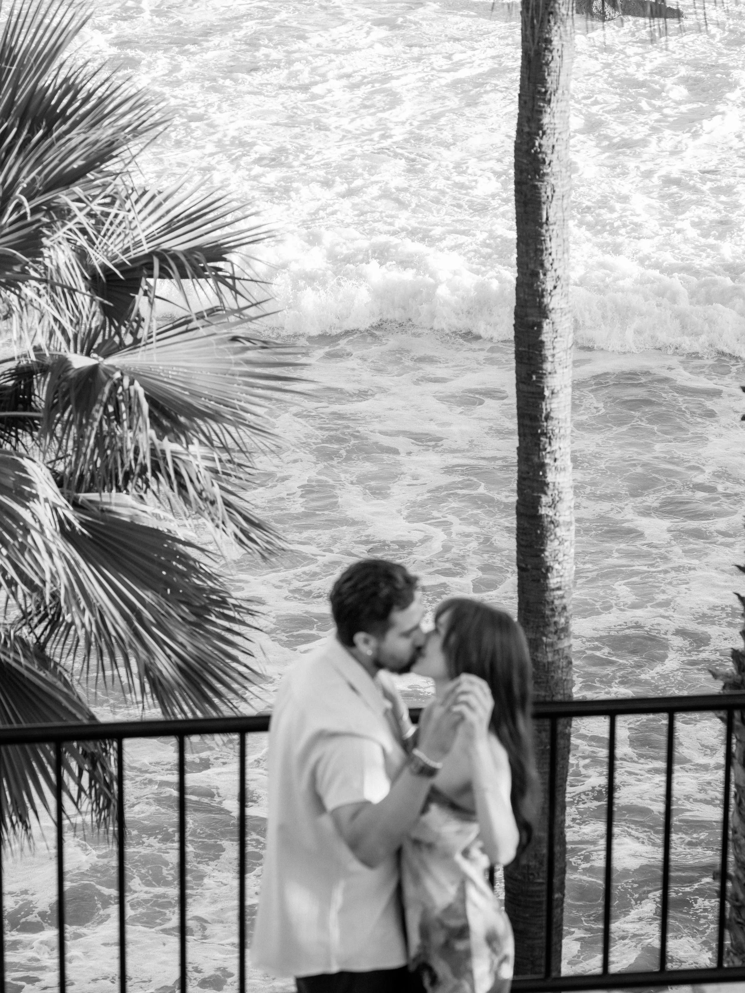 A couple kisses on a balcony overlooking the ocean, with palm trees in the foreground and waves crashing below.