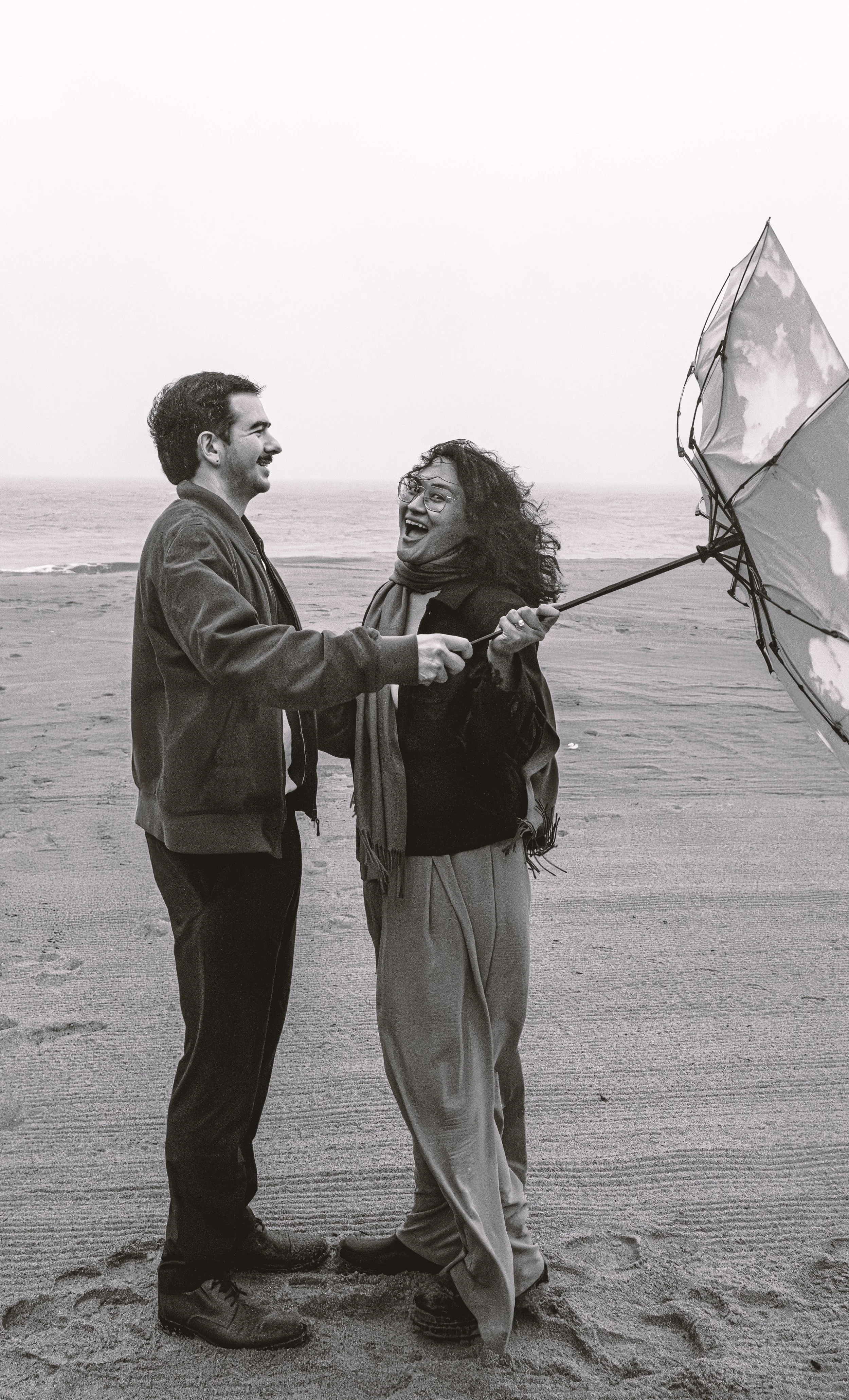 Two people, a man and a woman, stand on a beach facing each other, smiling and laughing. The woman holds an open umbrella with a cloud pattern, and the ocean can be seen in the background.