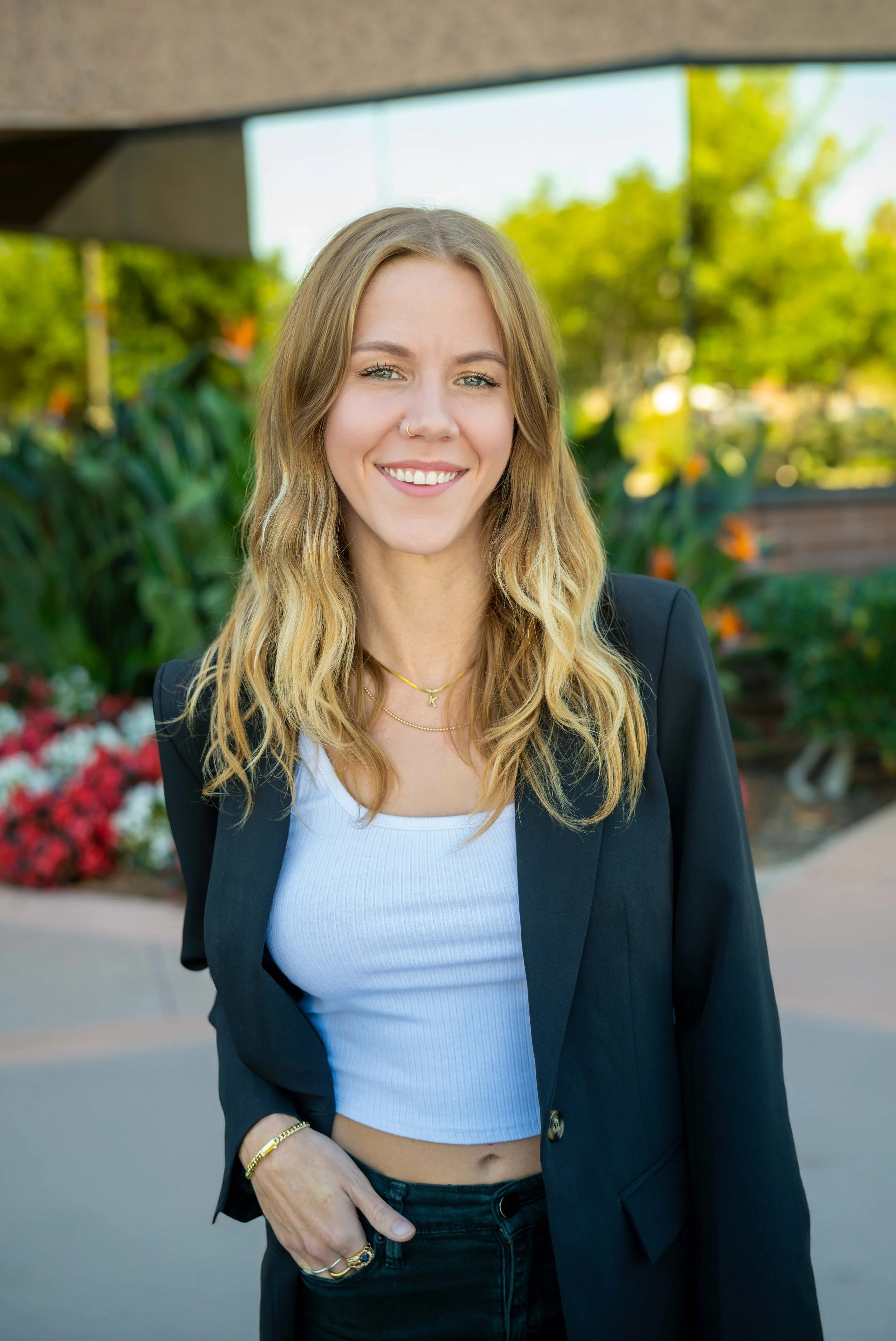 A young woman with long blond hair, wearing a black blazer over a white ribbed crop top and black jeans, smiling outdoors with greenery and flowers in the background.