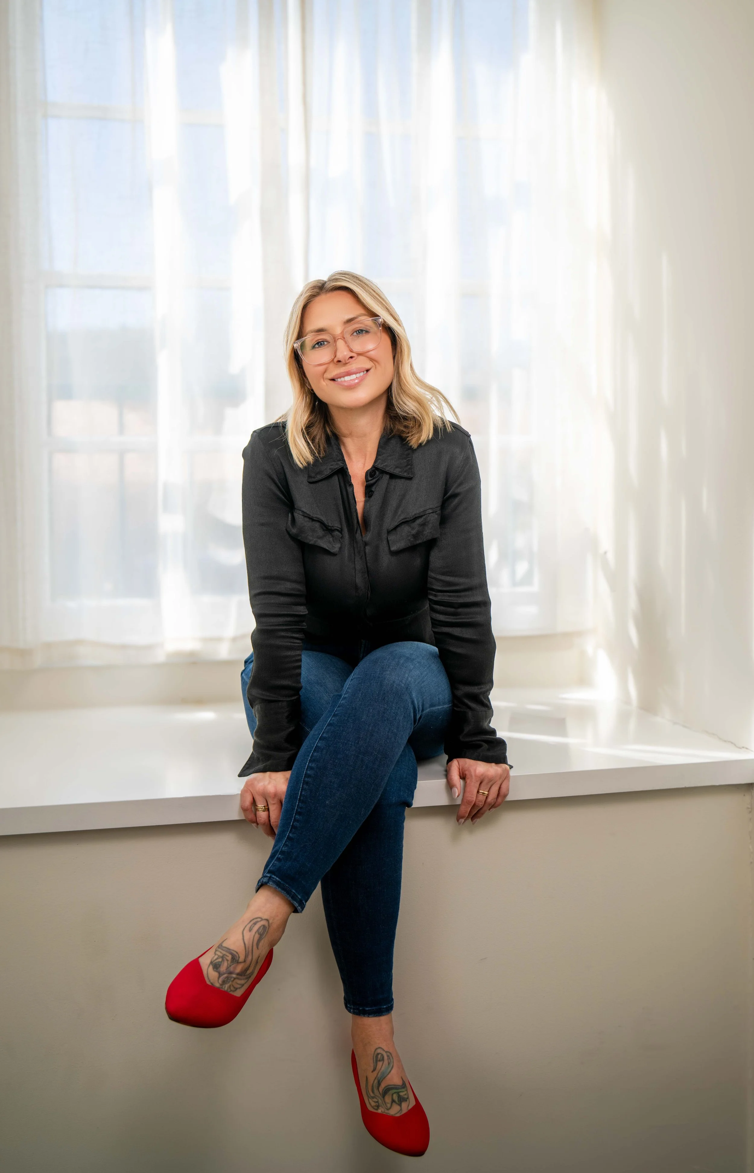 A woman with blonde hair and glasses sitting on a white ledge in front of a window with sheer curtains, crossing her legs with red high heels and tattoos on her feet, smiling.