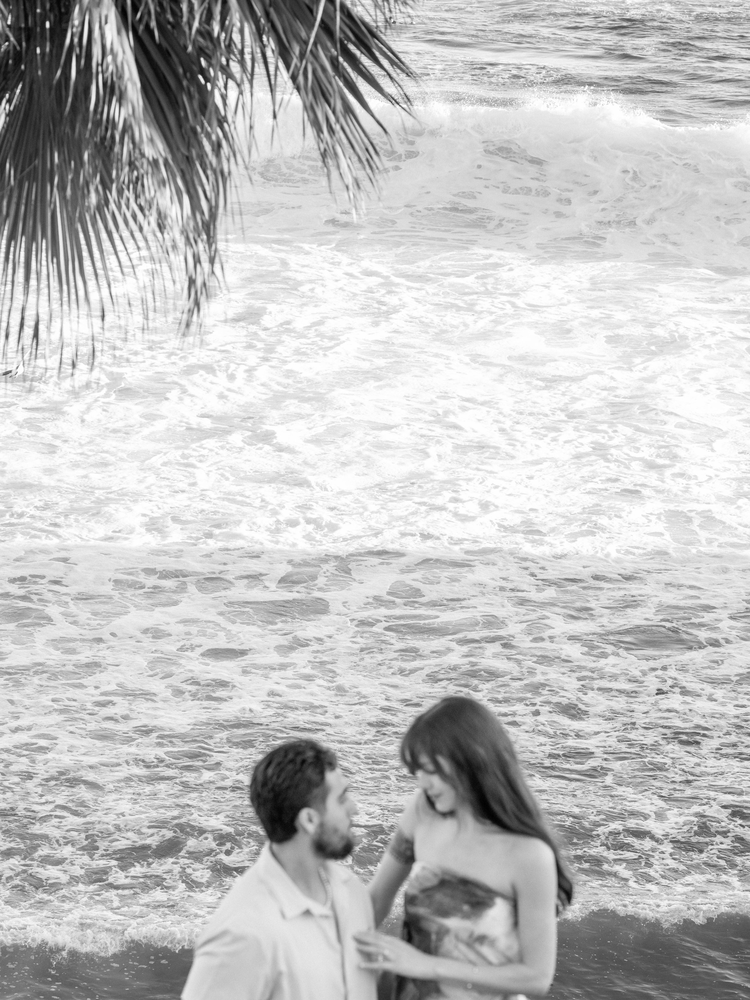 A black and white photo of a couple kissing on the beach, with the ocean and waves in the background and palm tree leaves hanging down from the top left corner.