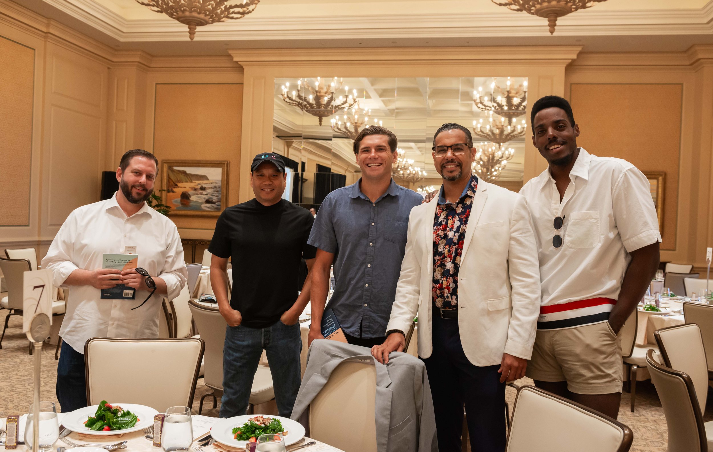 A group of five men standing together in a banquet hall, smiling at the camera. The hall has ornate chandeliers and framed artwork on the walls, with decorated tables and plates of food in the foreground.