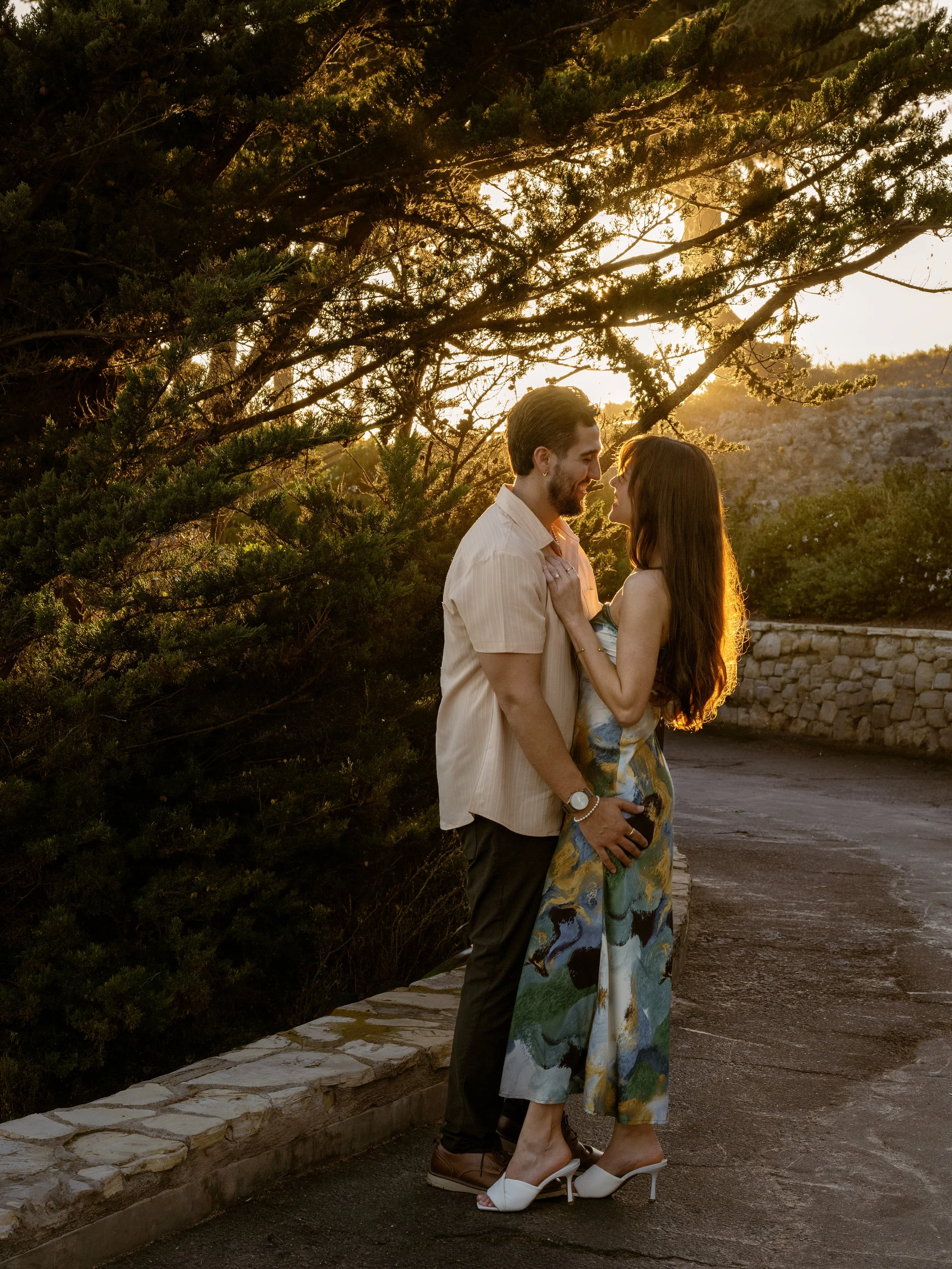 A couple stands close face-to-face on a paved path during sunset, with trees and a stone wall in the background, sharing a tender moment.