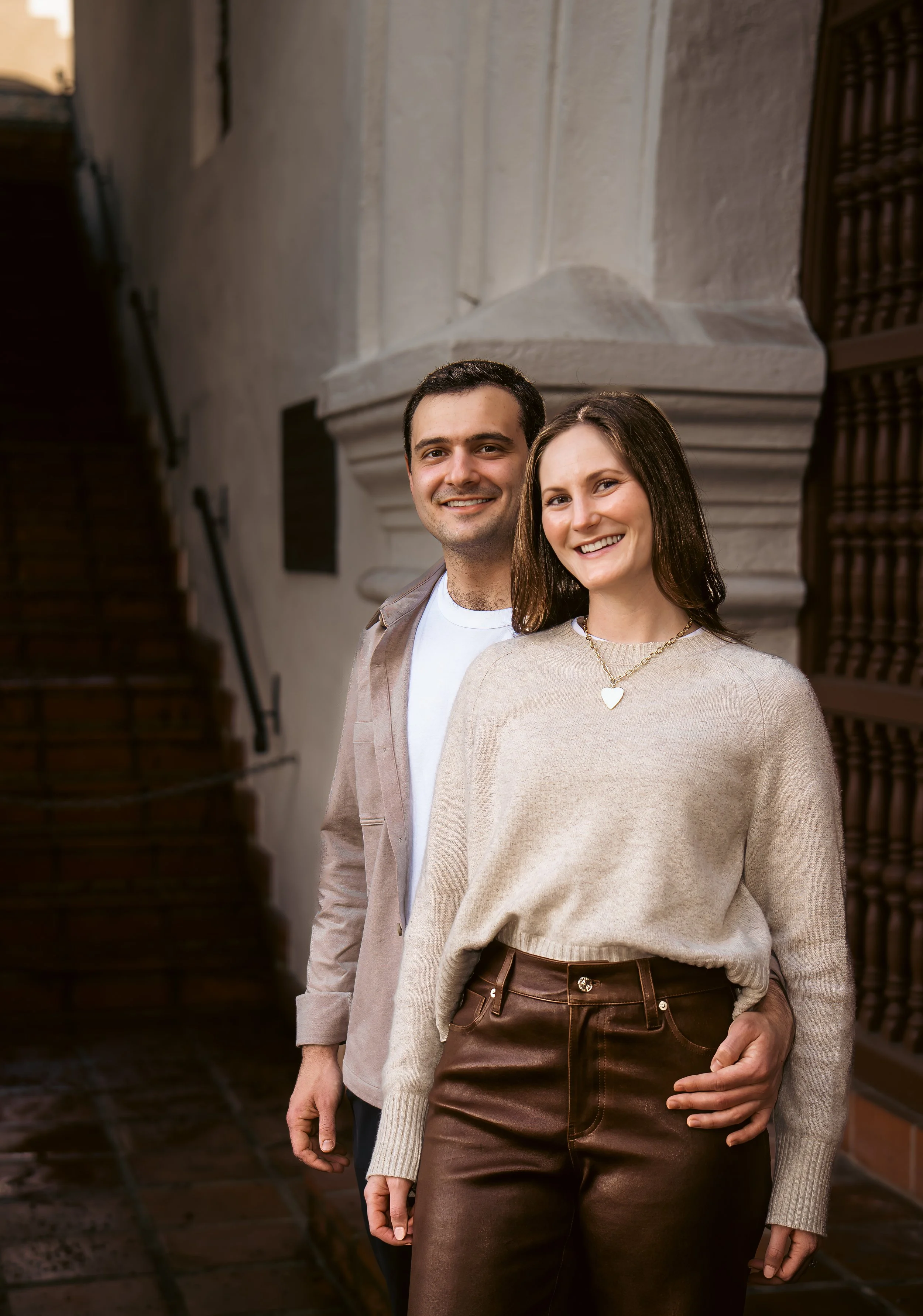 A smiling man and woman standing close together outside in front of a stone building, with dark stairs and wrought iron details.