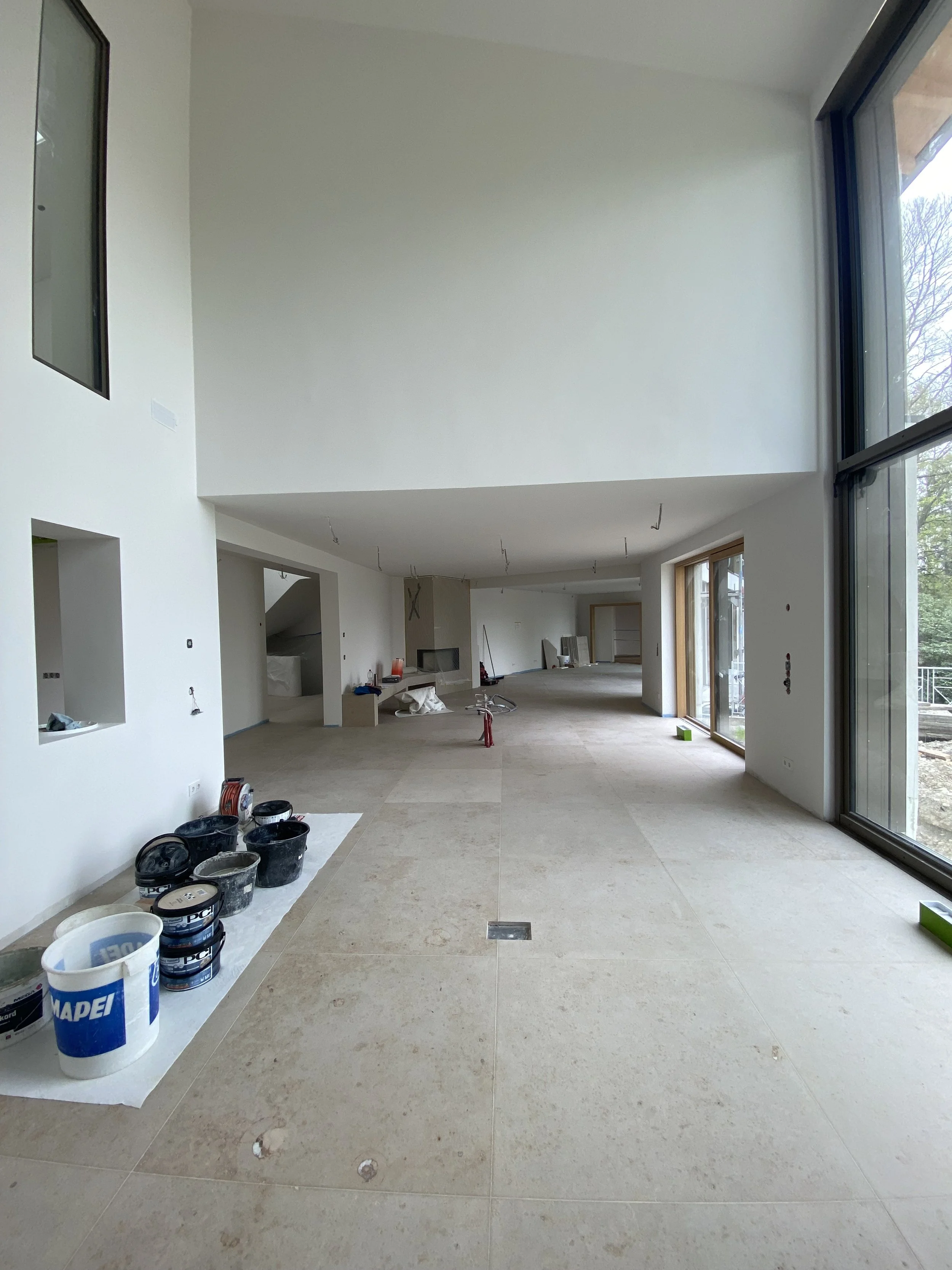 Unvollendeter Innenraum eines Hauses mit weißen Wänden und großen Fenstern, Bauutensilien und Materialien liegen auf dem Boden.