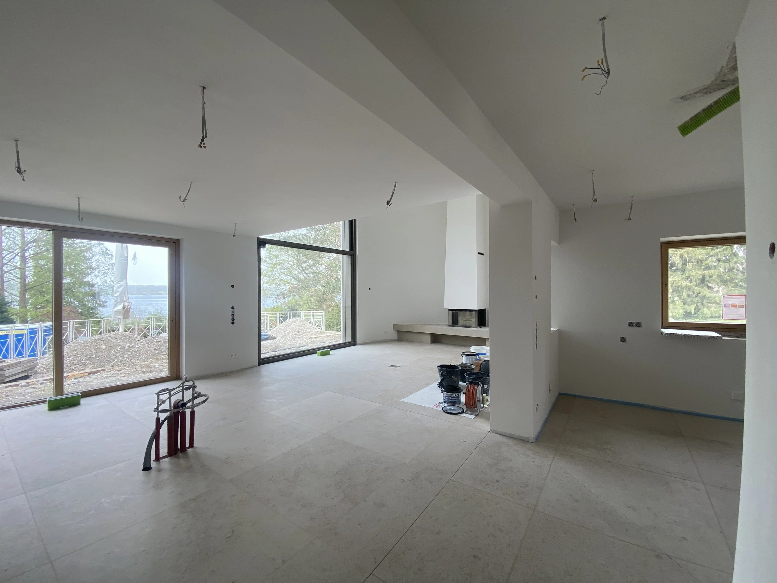 Innenansicht eines unfertigen Hauses mit großen Fenstern, weiß gestrichenen Wänden, offenem Grundriss, keine Möbel, Bauarbeiten sichtbar im Raum, Bauelemente und Werkzeuge auf dem Boden, Blick nach draußen auf eine Bau- und Naturlandschaft.