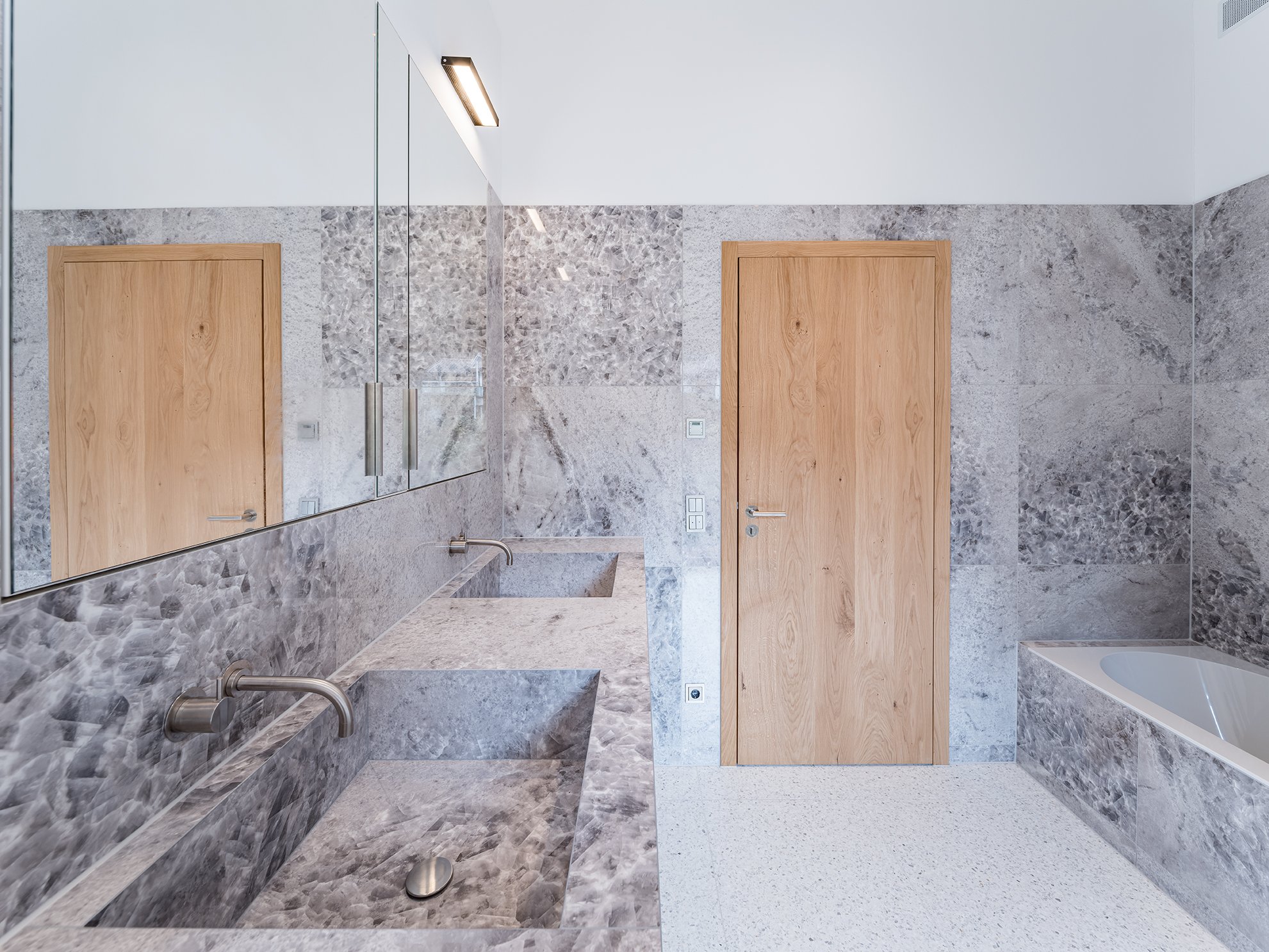 Moderner Badezimmer mit grauer Marmorwand und -abdeckung, Holz-Türen, großem Spiegel, Waschbecken, Wasserhahn, Badewanne, flachem Bodenbelag.