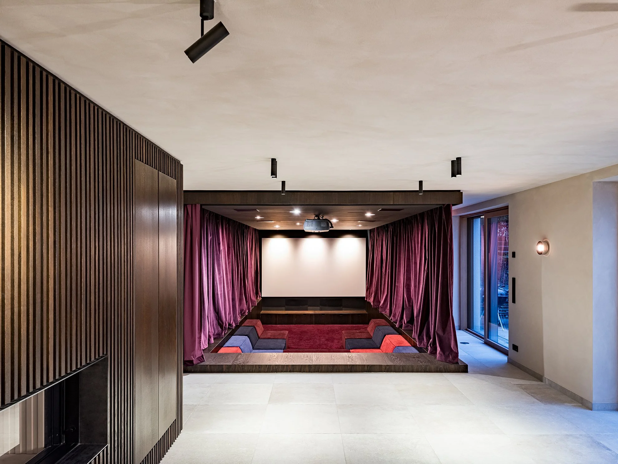 Ein Heimkinoraum mit einer großen weißen Leinwand, umrahmt von violetten Vorhängen, einem kleinen Sitzbereich mit rot-schwarz gepolsterten Sitzbänken und einem dunklen Holzboden.