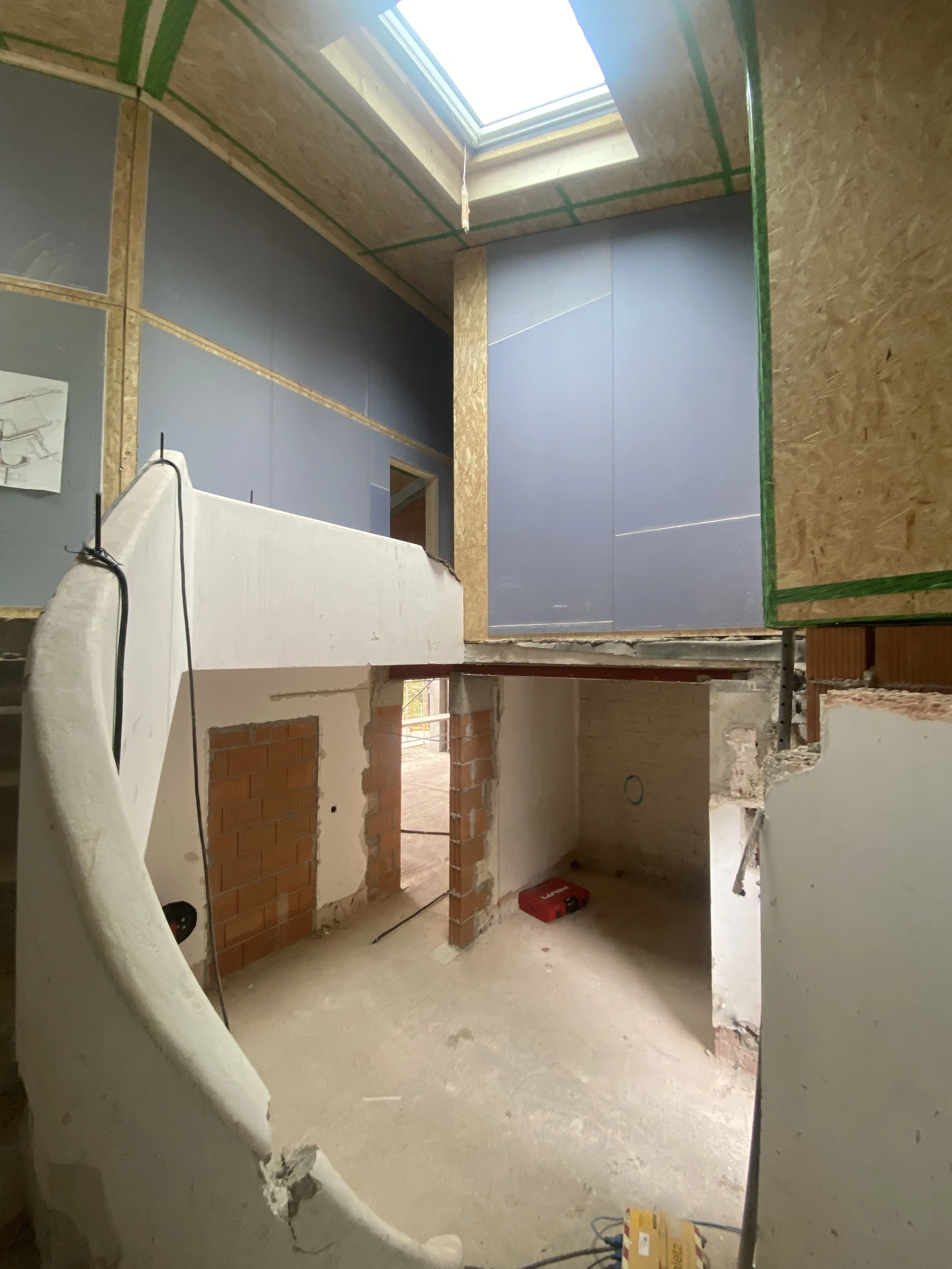 Innenaufnahme eines Bauprojekts mit unvollständigen Wänden, einem Dachfenster, das Tageslicht in den Raum lässt, und Baumaterialien auf dem Boden.