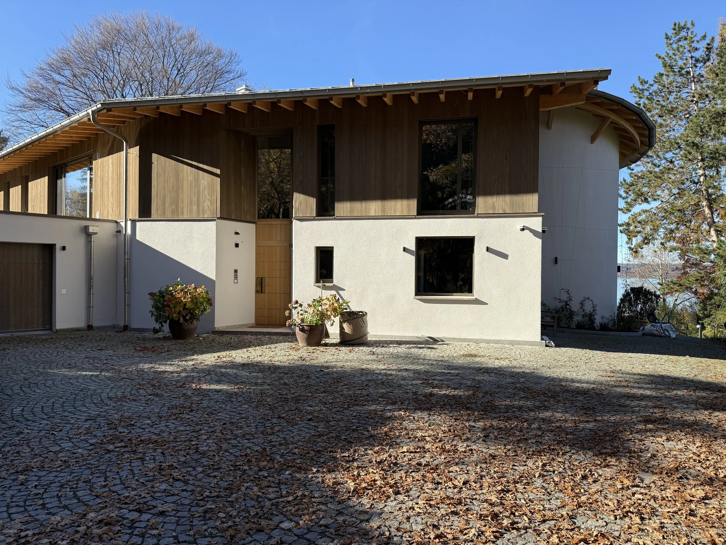 Modernes zweistöckiges Haus mit Holz und weißem Putz, große Fenster, Holz-Tür, zwei große Pflanzkübel mit Pflanzen, gepflasterter Hof, Bäume im Hintergrund, klarer blauer Himmel.