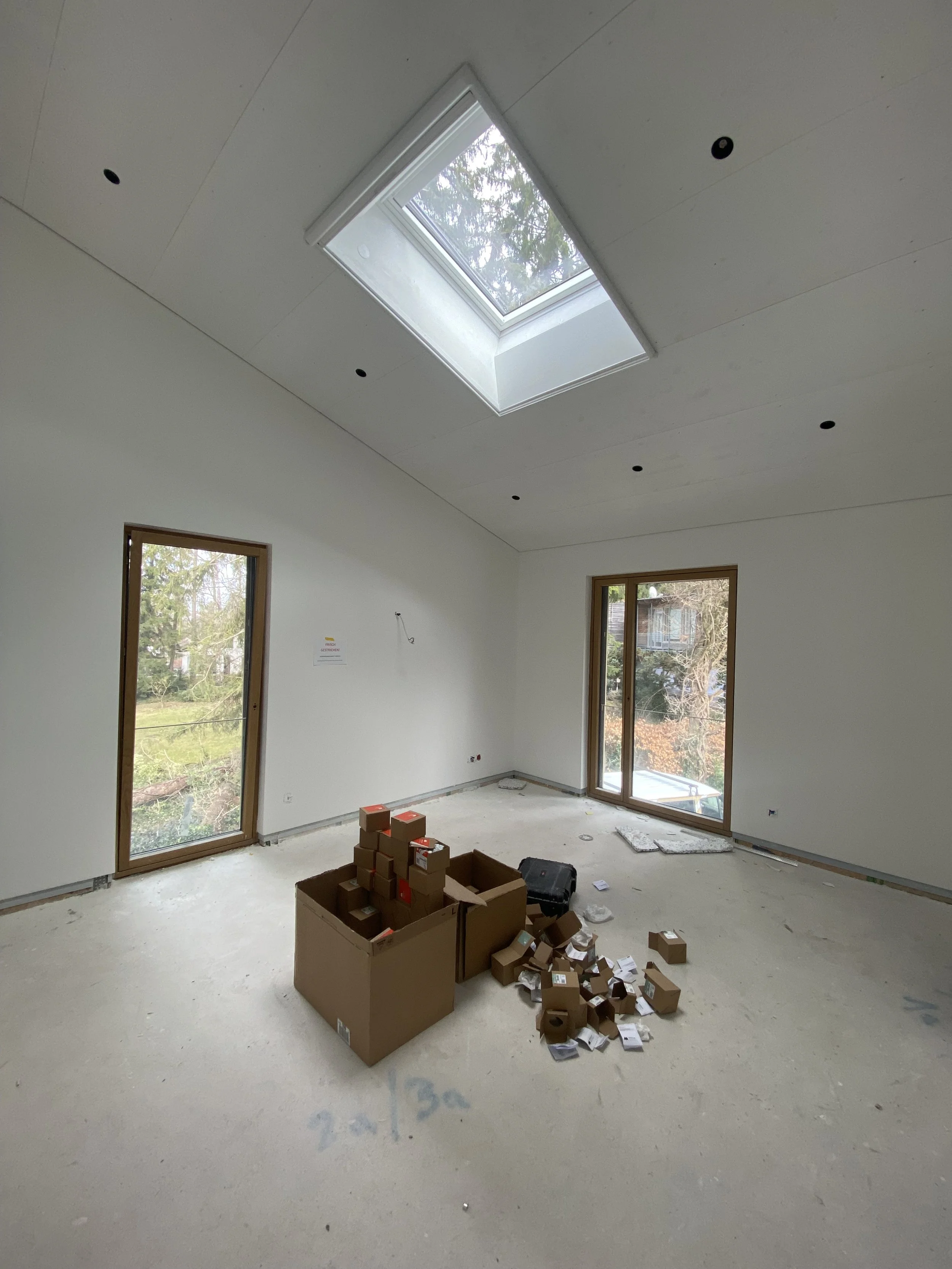 Ein leerer Raum mit weißen Wänden und großen Fenstern. Das Dach hat eine Dachgaube mit einem Fenster. Es befindet sich Baumaterial und Verpackung auf dem Boden.