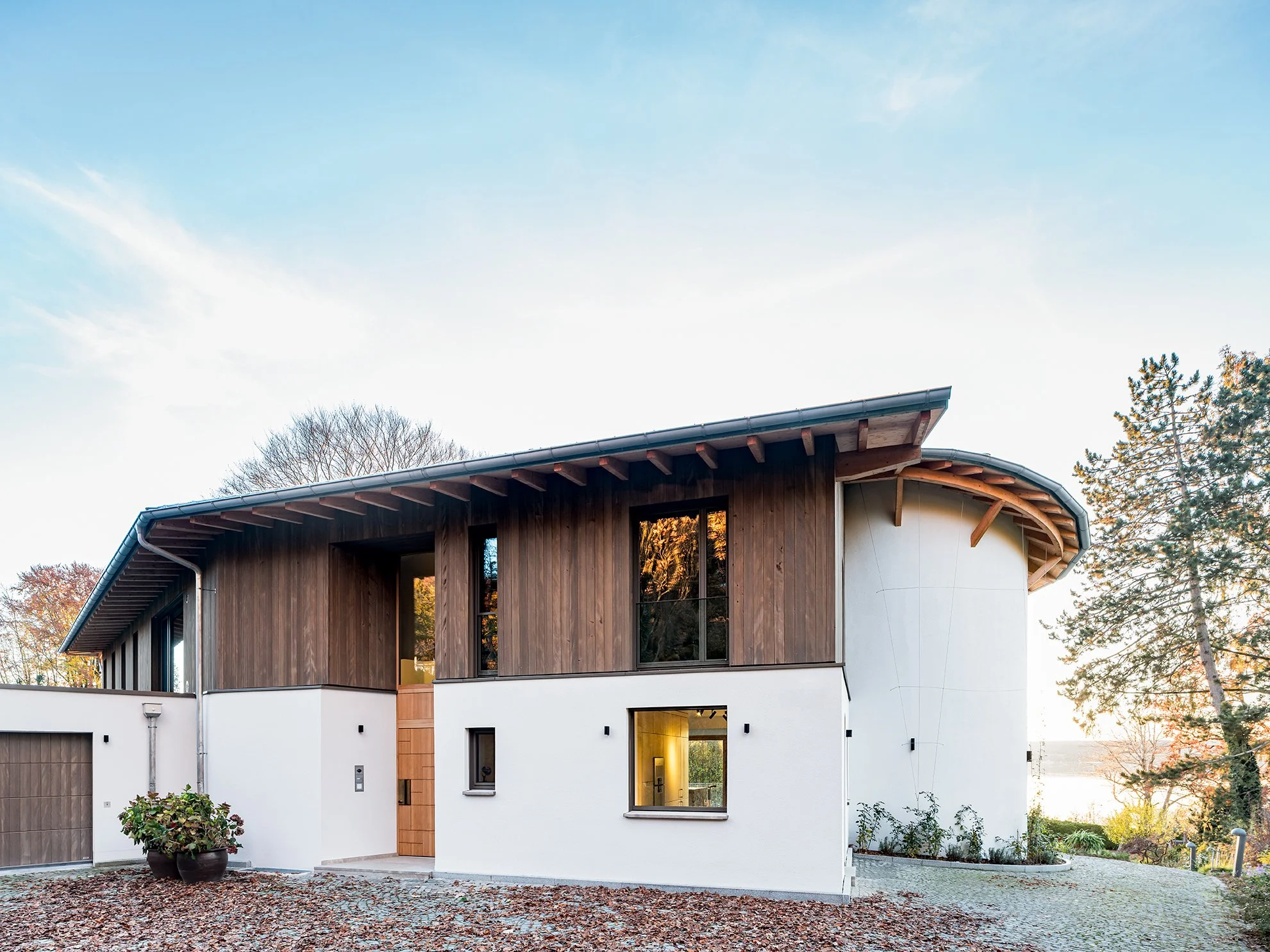 Modernes Haus mit Holzfassade und weißen Wänden, große Fenster, umgeben von Bäumen, bei Sonnenuntergang