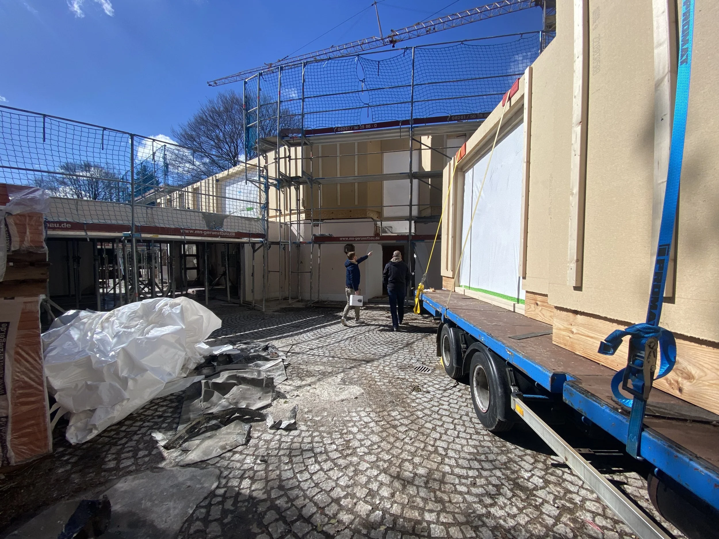 Baustelle mit Baugerüst und einem Bauwagen, zwei Personen im Gespräch, Materialien auf dem Boden, sonniger Tag, Straßenpflaster, Baum im Hintergrund.