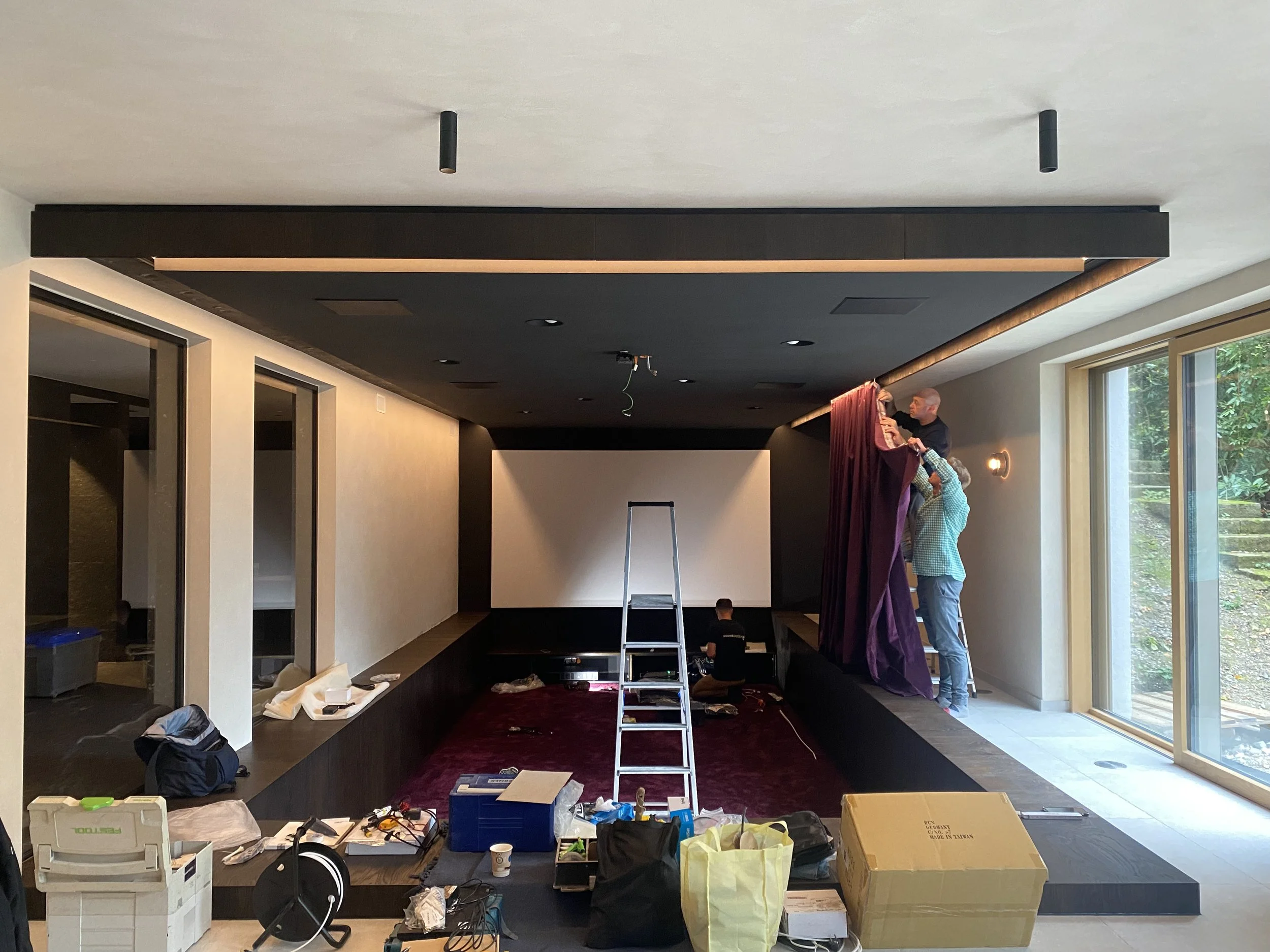 Zwei Männer hängen einen Vorhang an die Decke eines Kinos, während sie auf einer Leiter stehen. Im Raum sind viele Werkzeuge und Pakete auf dem Boden verstreut.