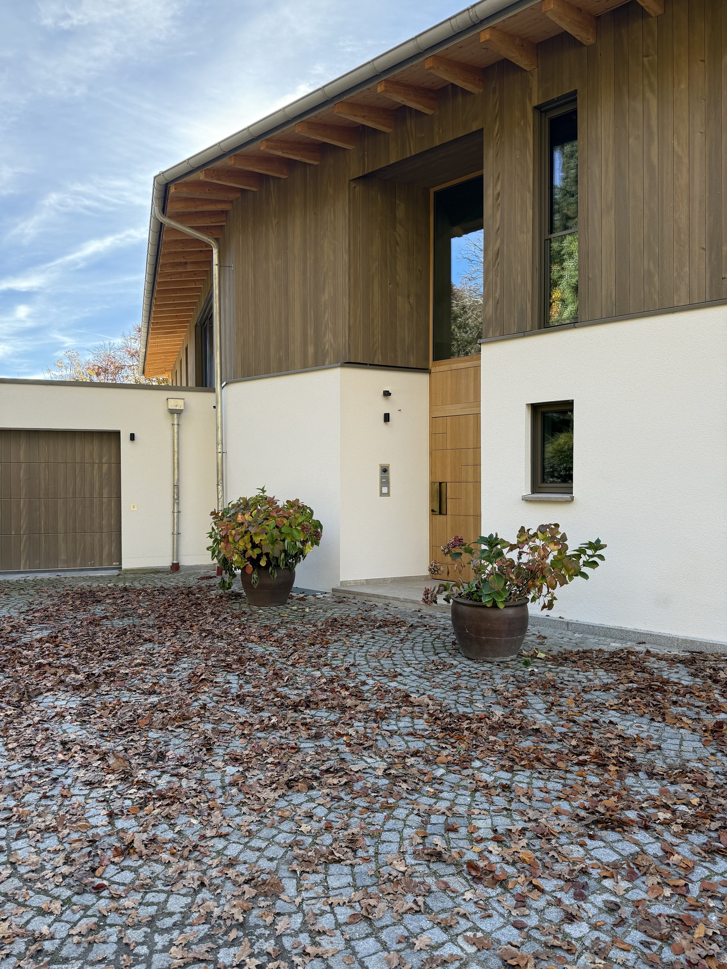 Modernes Haus mit Holz- und Putzfassade, zwei große Pflanzkübel mit Pflanzen vor dem Eingang, Laub auf gepflastertem Weg, blauer Himmel mit wenigen Wolken.