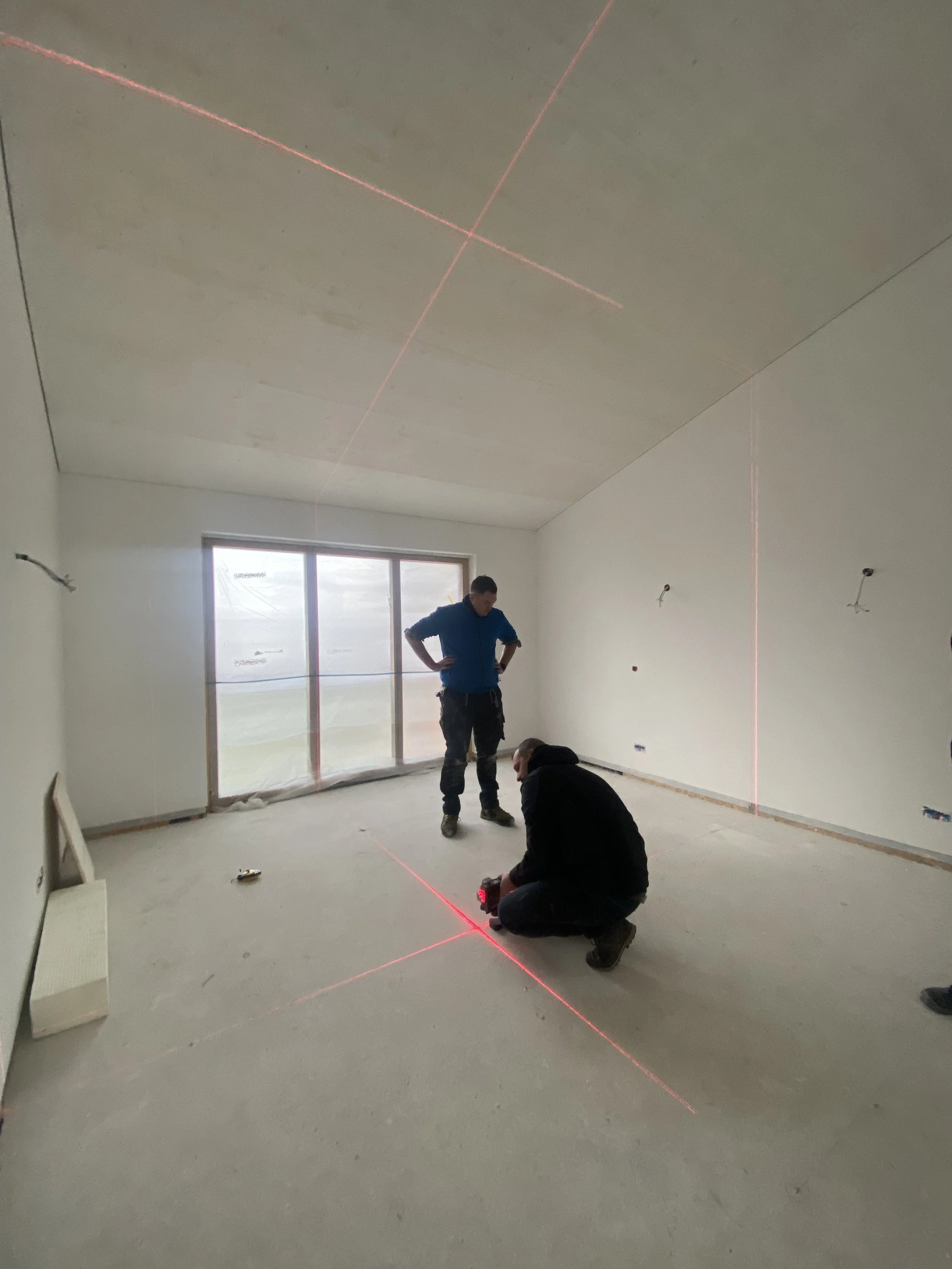 Zwei Arbeiter vermessen einen Raum mit roten Laserlinien, in einem hellen Raum mit großen Fenstern.