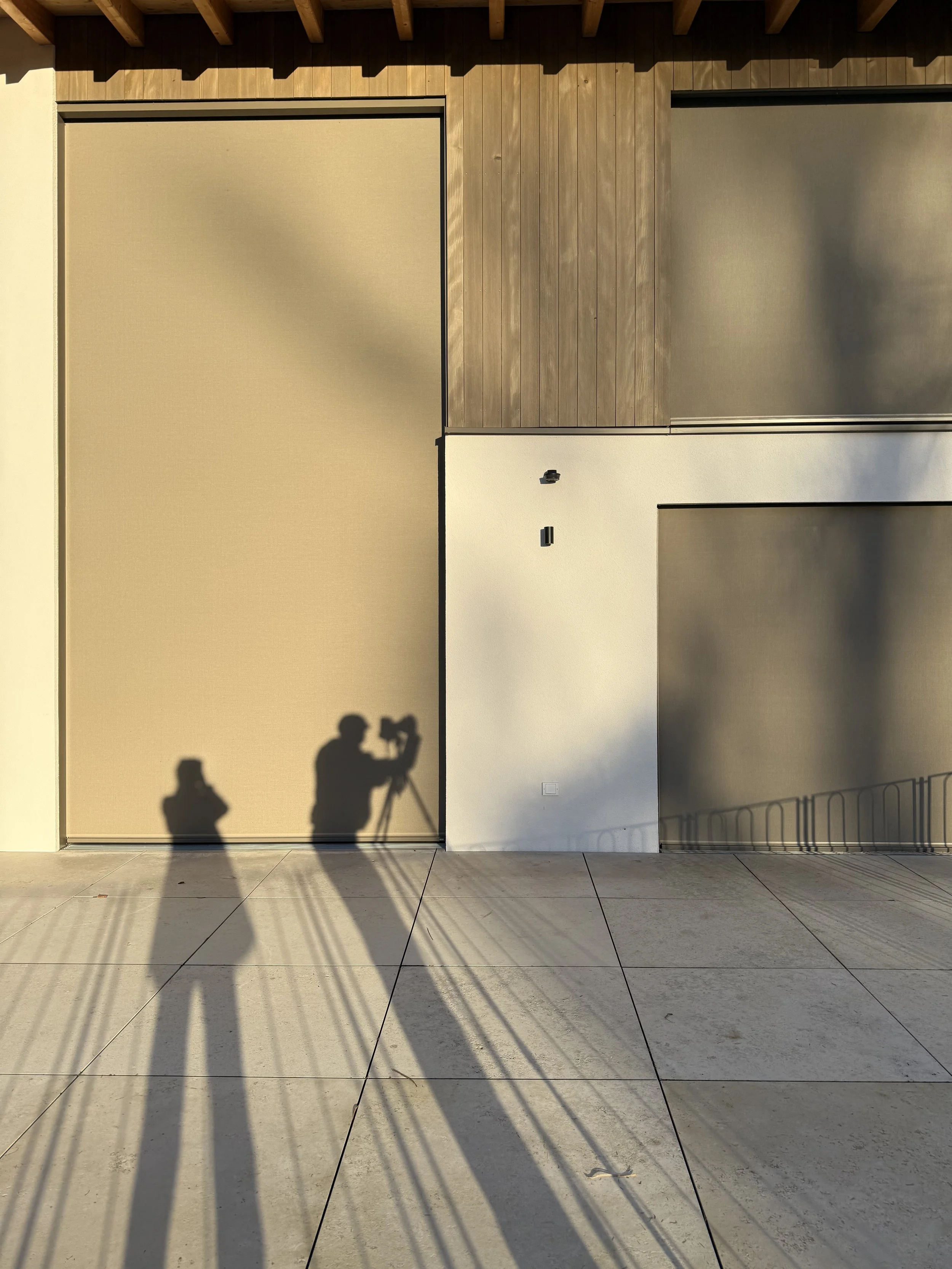 Schatten von zwei Personen, einer mit Kamera, auf einer Gebäudefront mit Holz- und Putzflächen bei Sonnenuntergang.