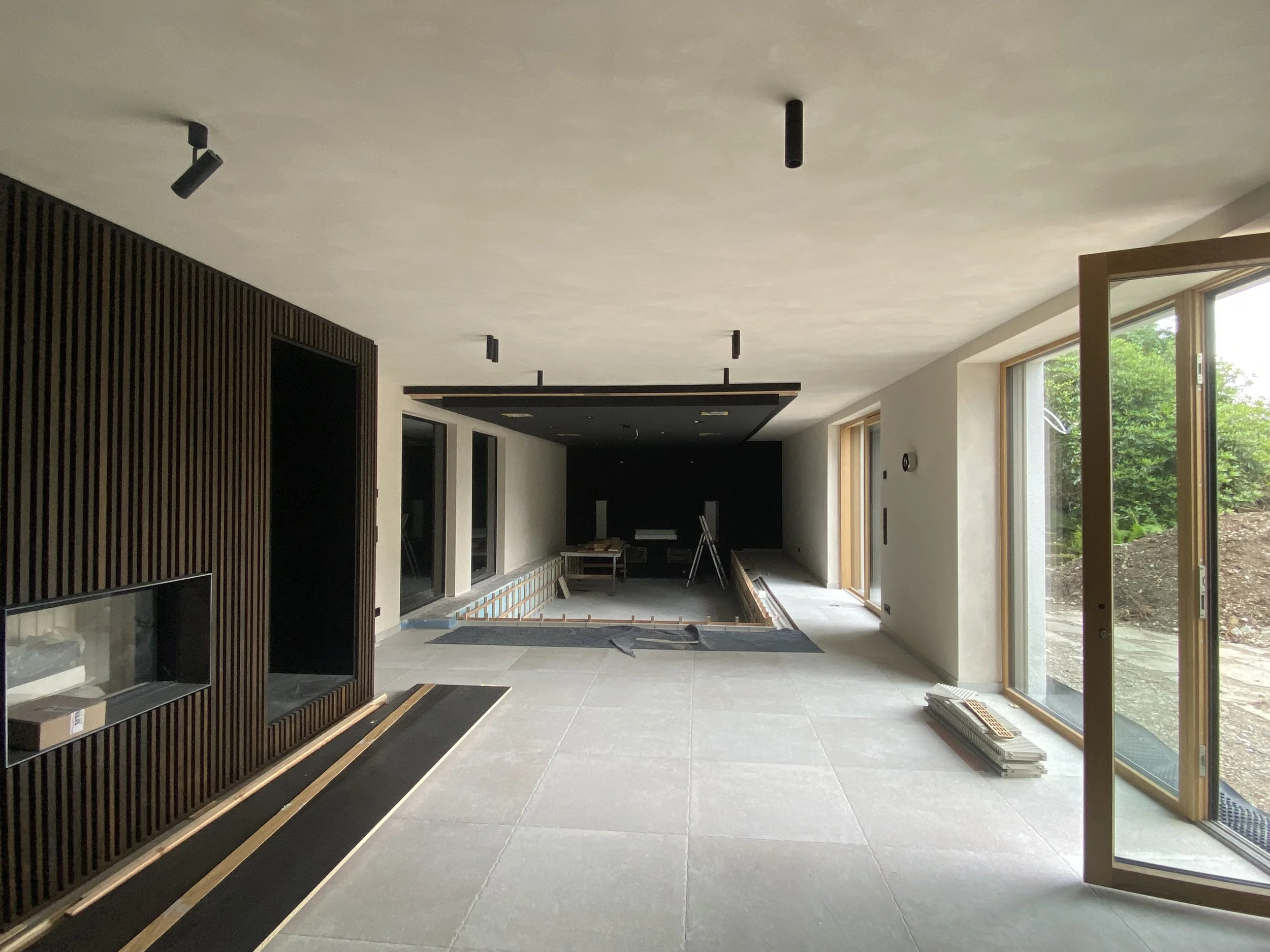 Innenraum eines Hauses unter Bau: unvollendete Treppe, Holzfenster, schwarze und weiße Wände, teilweise fertig verlegte Fliesen, Bauwerkzeuge, sichtbarer Garten im Hintergrund