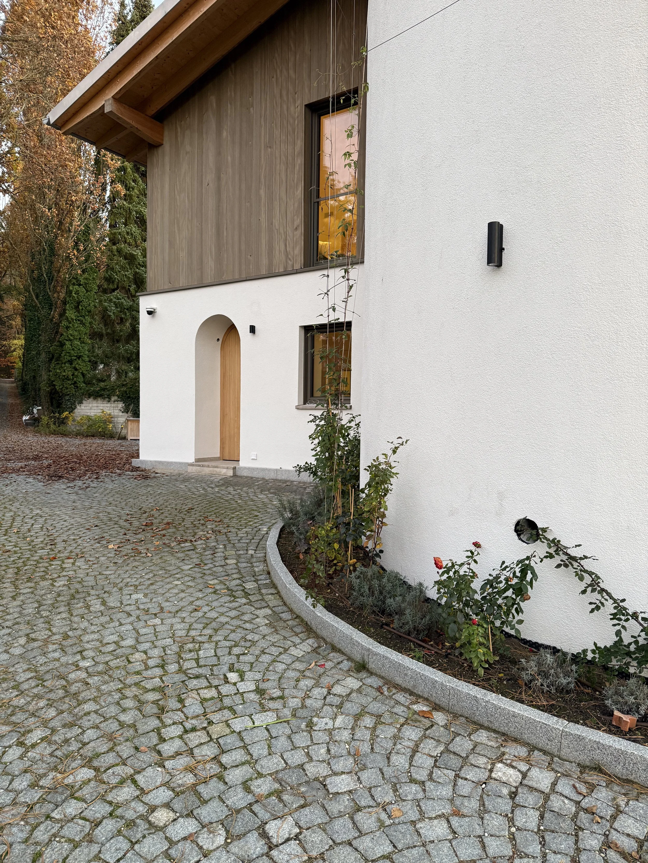 Modernes Haus mit weißer und holziger Fassade, Eingang mit Bogen, Pflanzbeet mit Rosen und anderen Pflanzen, gepflasterter Weg aus Cobblestones, umgeben von Bäumen im Herbst.