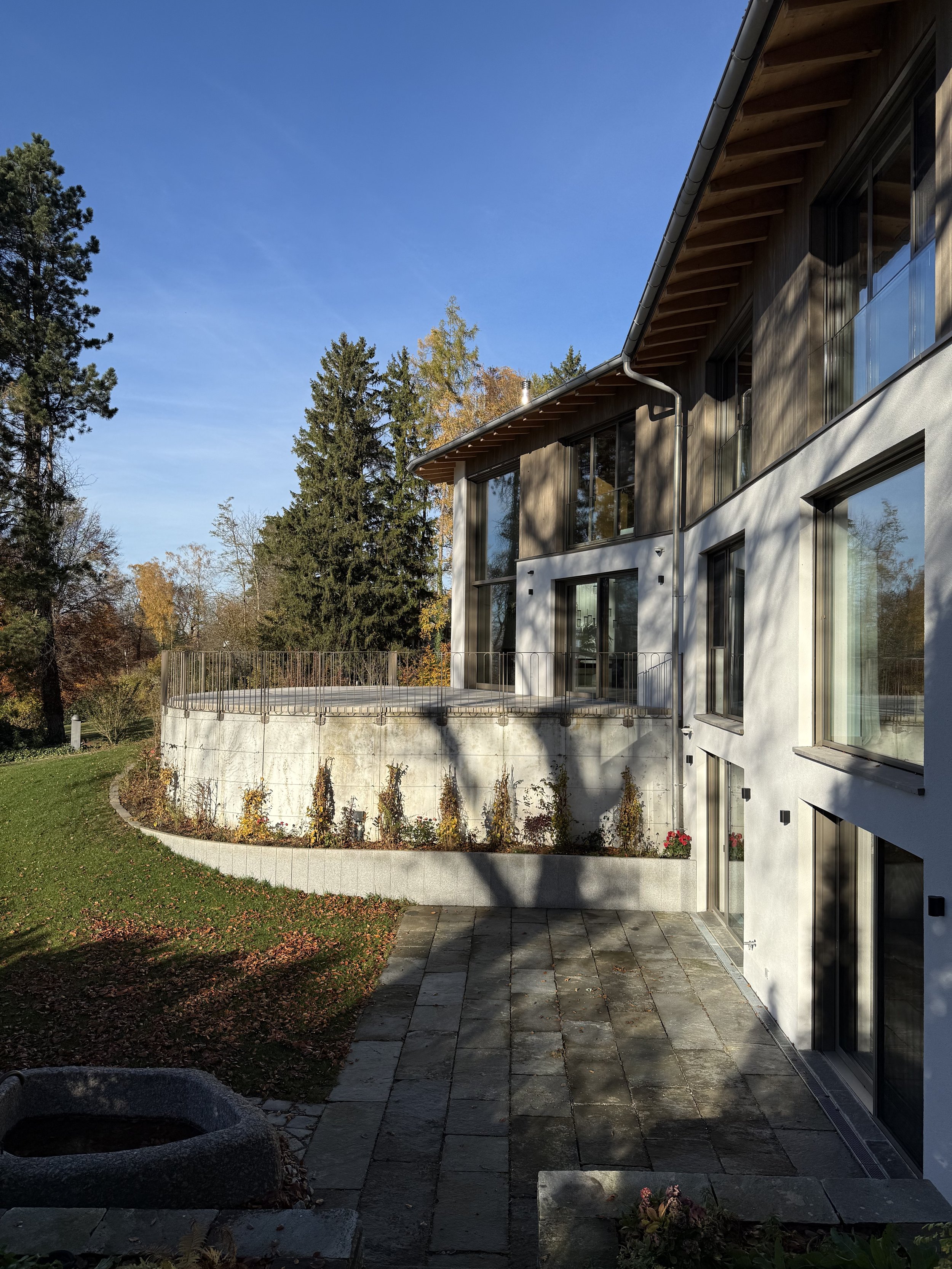 Modernes Haus mit großen Fenstern und Terrasse, umgeben von Bäumen und Garten, bei sonnigem Wetter.