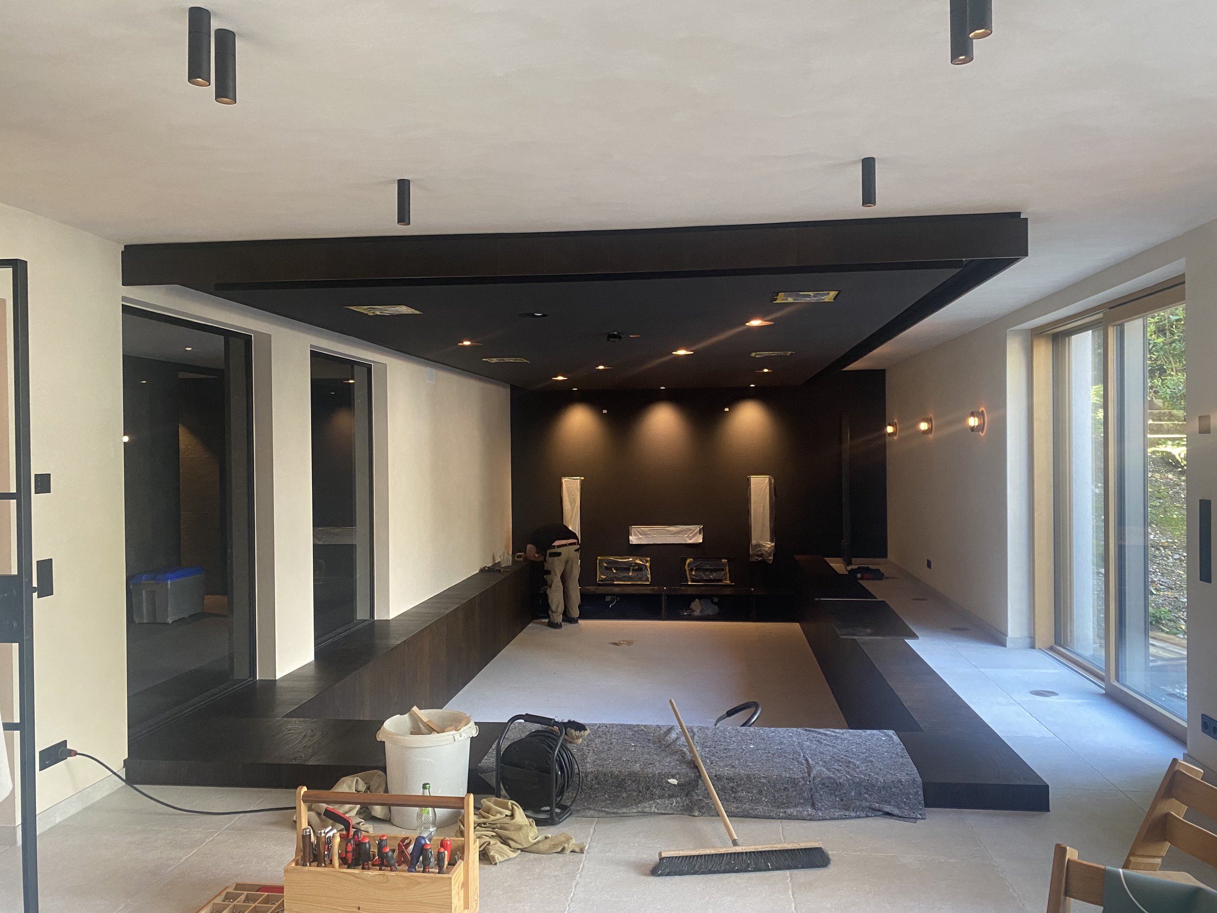 Innenaufnahme eines Hauses im Bau mit dunklem Fußboden und weißen Wänden, große Glasfenster, Steinelemente an der Wand, Bauwerkzeug und Materialien im Raum, Person arbeitet an der Wand.