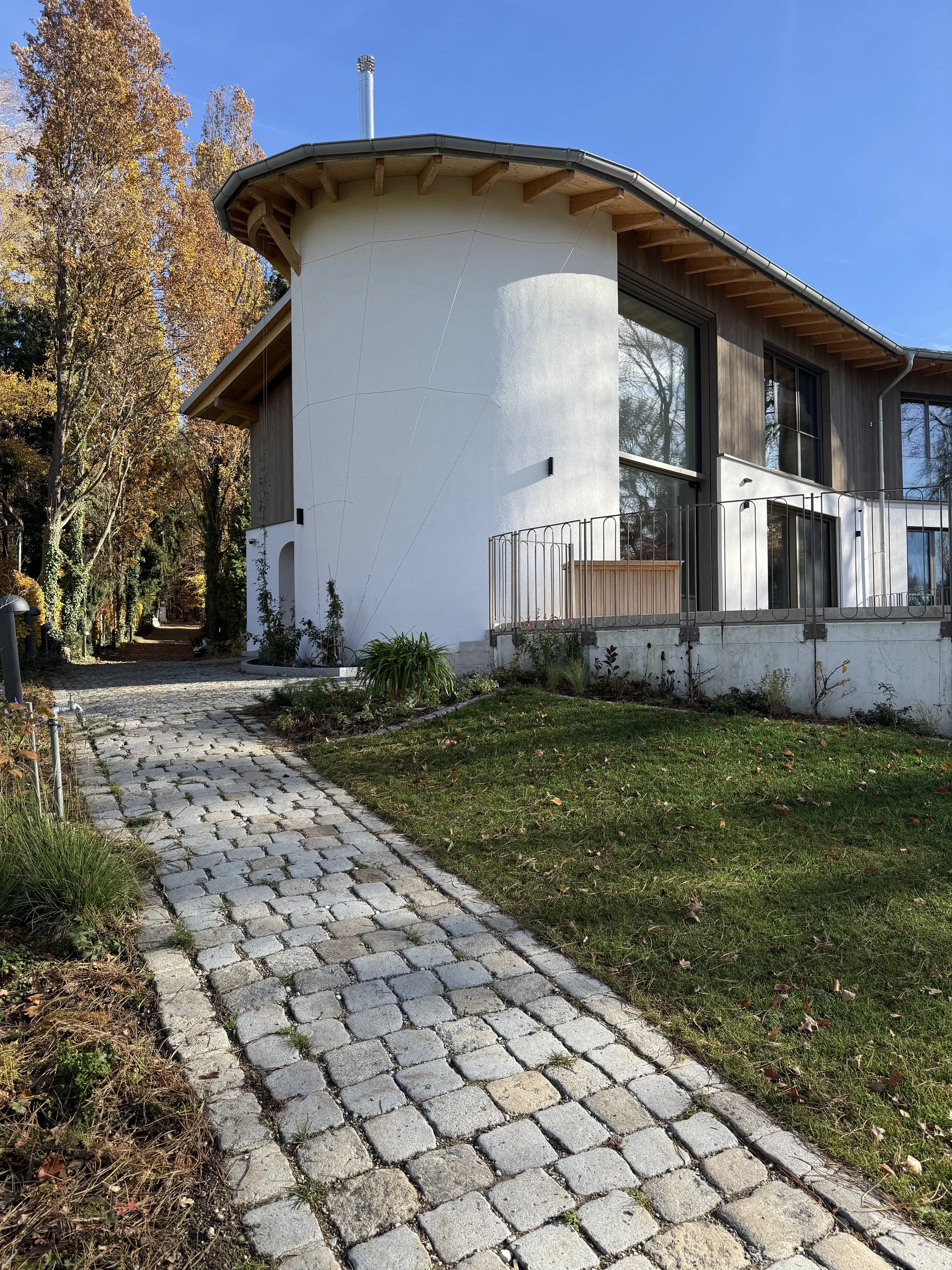 Modernes Haus mit einer halb gewölbten weißen Wand, große Fenster und umgeben von einem gepflegten Garten und einem gepflasterten Weg, im Herbst bei blauem Himmel.