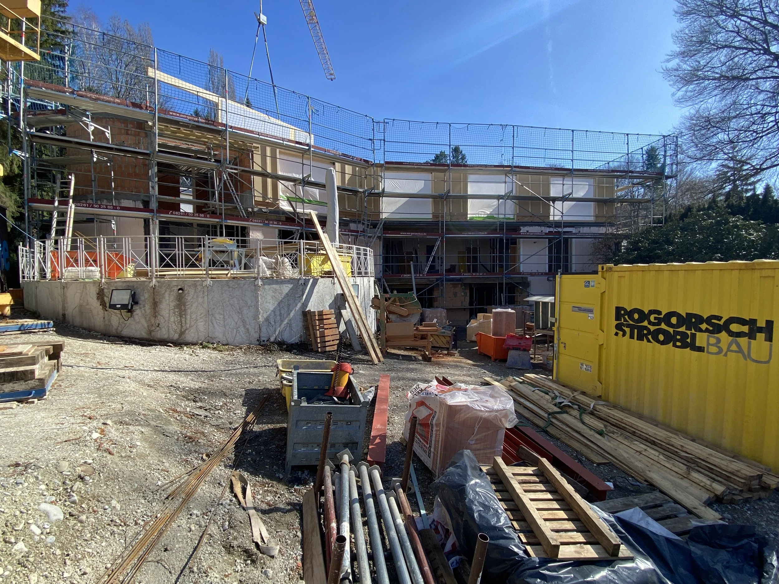 Baustelle mit Gerüsten, Baumaterialien und Baucontainer, im Hintergrund ein Gebäude im Bau, bei strahlendem blauen Himmel.
