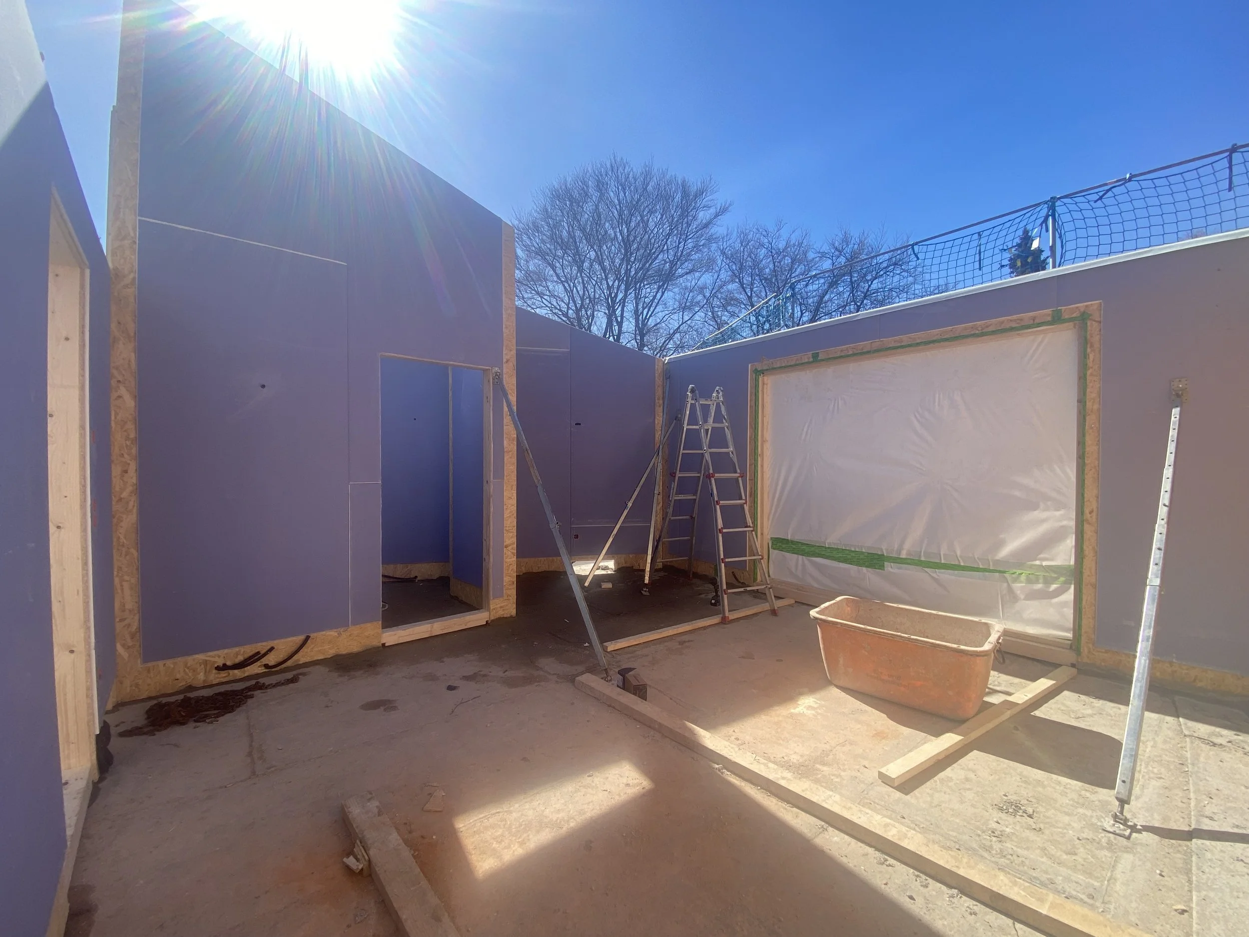 Baustelle im Innenraum eines Hauses mit blauen Wandplatten, Leiter und Baumaterialien, sonniger Himmel im Hintergrund.