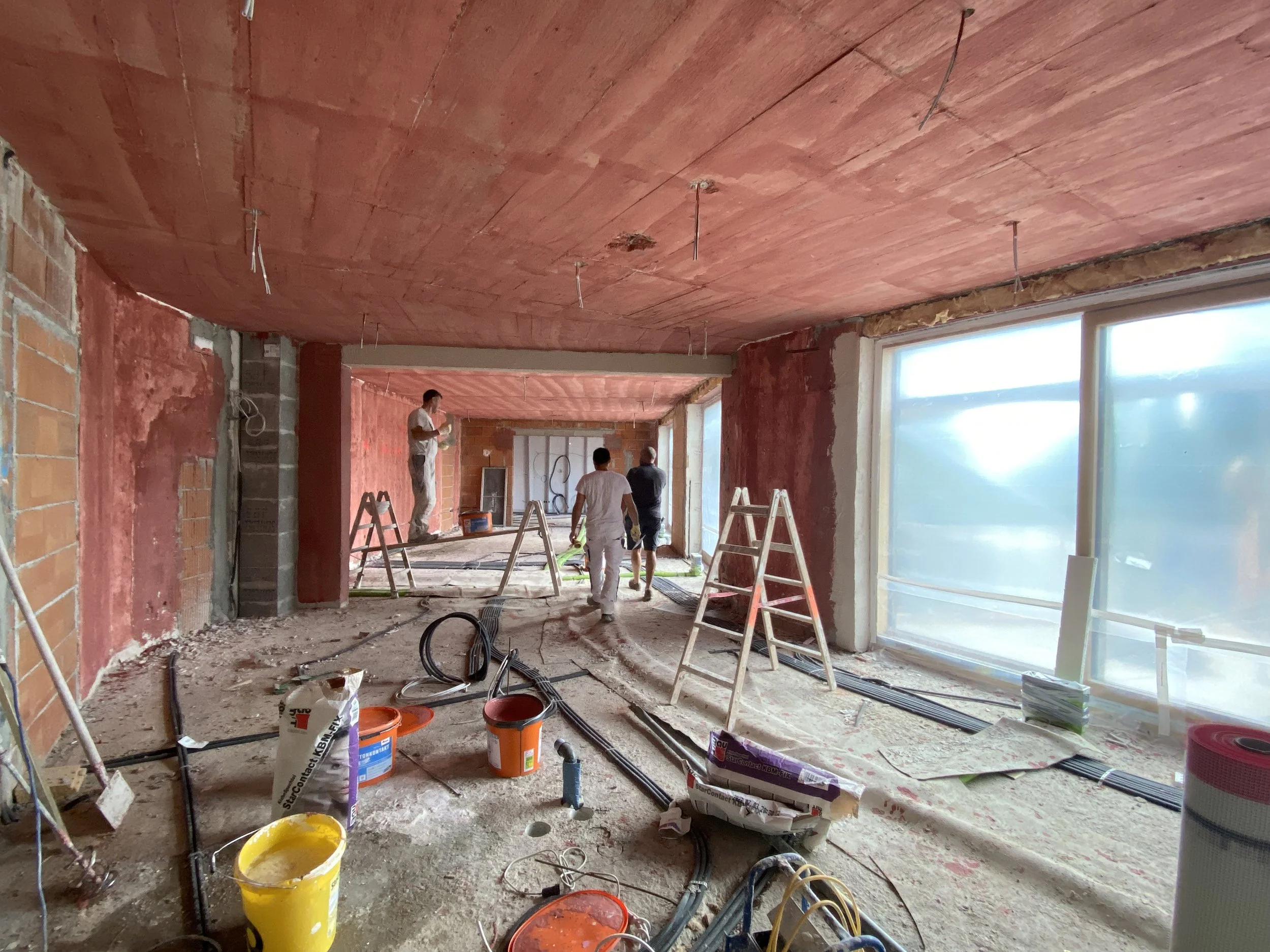 Innenraum eines Gebäudes im Bau, mit drei Menschen, die an der Decke und am Boden arbeiten, Bauwerkzeuge, Kabel und Baumaterial auf dem Boden, große Fenster mit Sicht nach draußen.