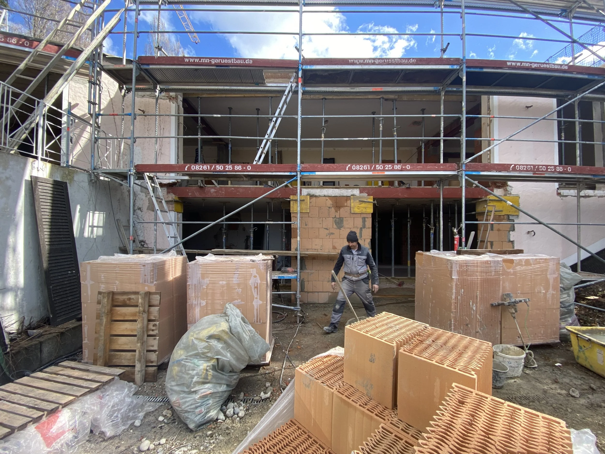 Baustelle mit Bauarbeitern, Gerüsten und Baumaterialien vor einem mehrstöckigen Gebäude. Ein Arbeiter steht mit einem Werkzeug in der Hand, im Hintergrund ist blauer Himmel mit Wolken sichtbar.