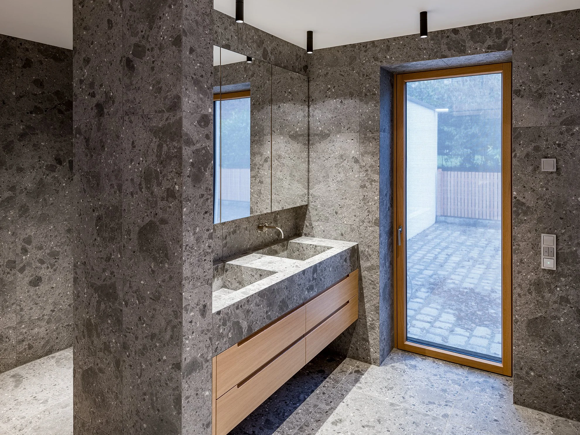 Moderner Badezimmerraum mit grauer Steinwand, waschbecken, großem Spiegel und glastür nach draußen.