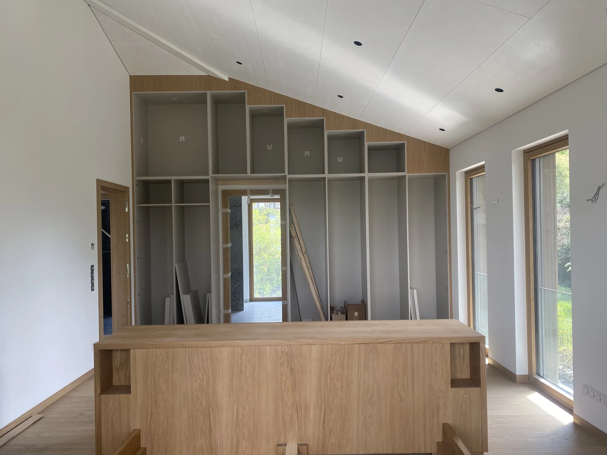 Innenansicht eines noch im Bau befindlichen Zimmers mit großen Fenstern, einer Holzbar, einer offenen Holzregal-Wand und weiß gestrichenen Wänden.