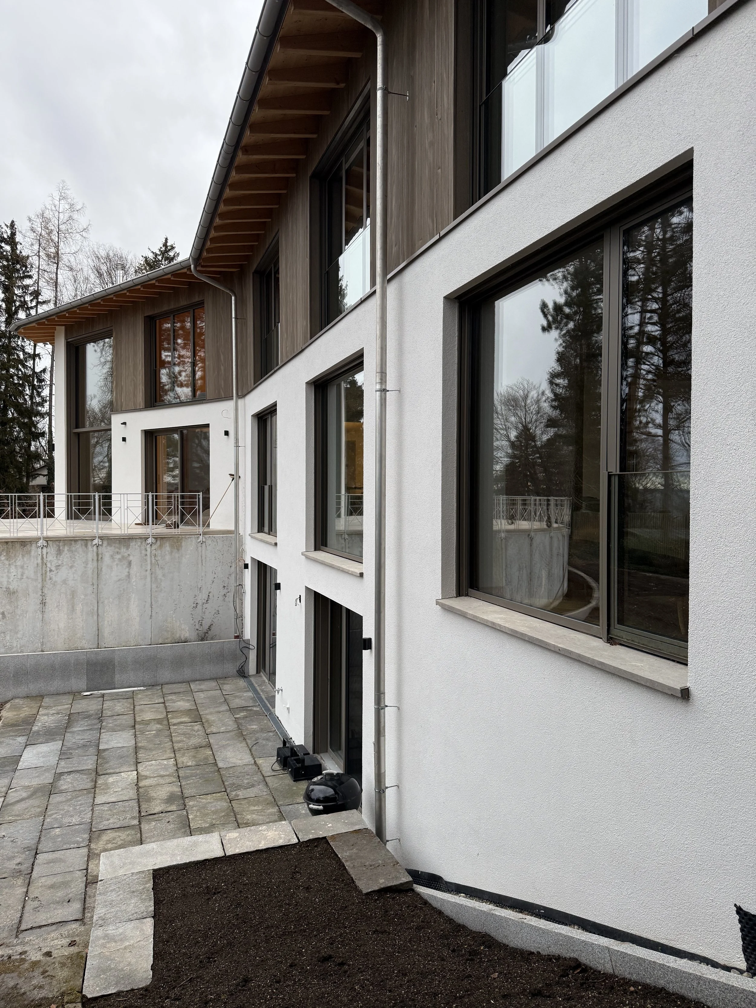 Moderne vierstöckige Hausfassade mit weißen und holzverkleideten Flächen, großen Fenstern und einer Terrasse im Hintergrund, umgeben von Bäumen.