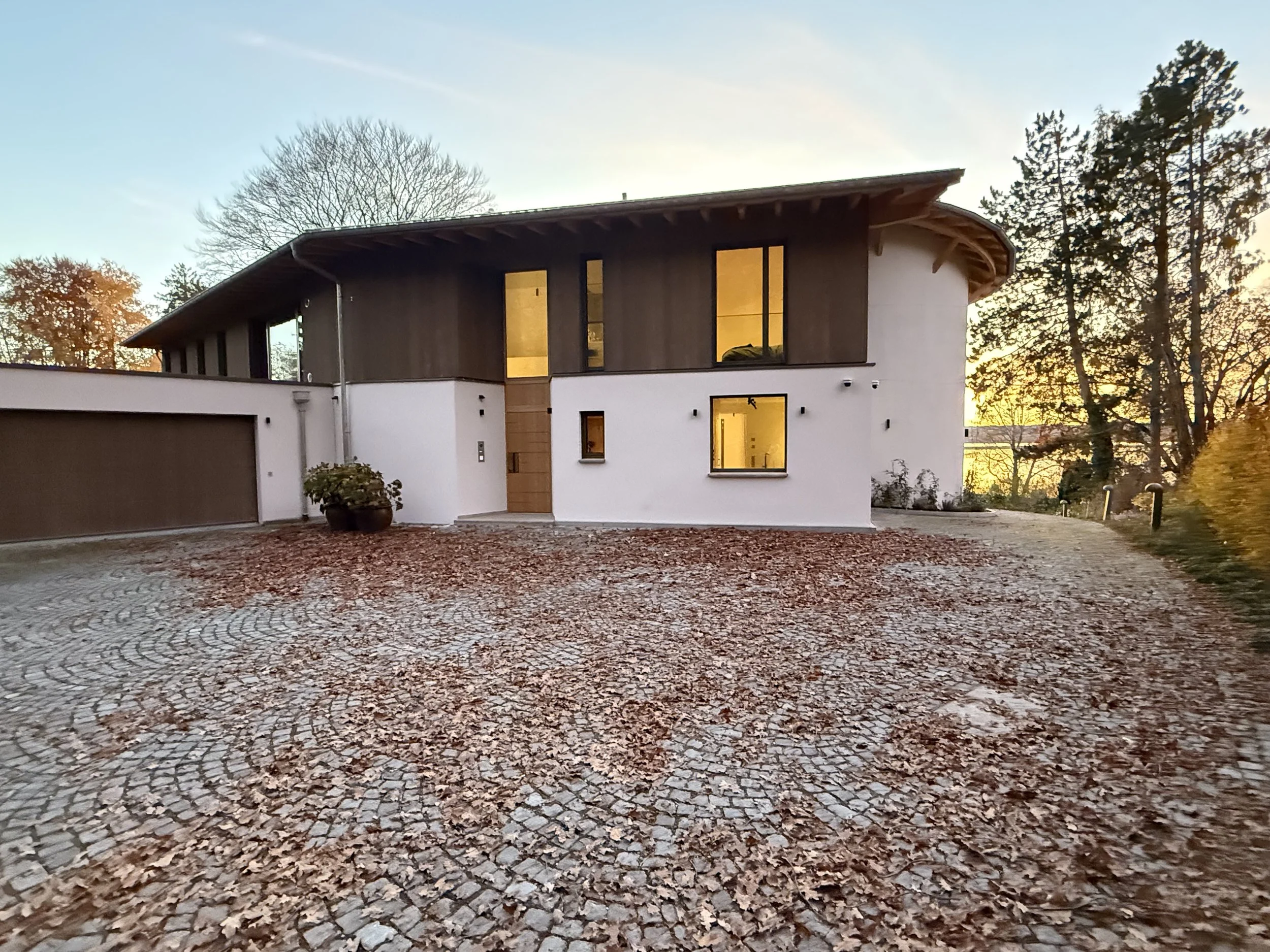 Modernes zweistöckiges Haus mit weißen und dunklen Holzwänden, große Fenster, eingezäunter Eingangsbereich, Herbstlaub auf gepflastertem Hof, umgeben von Bäumen bei Sonnenuntergang.