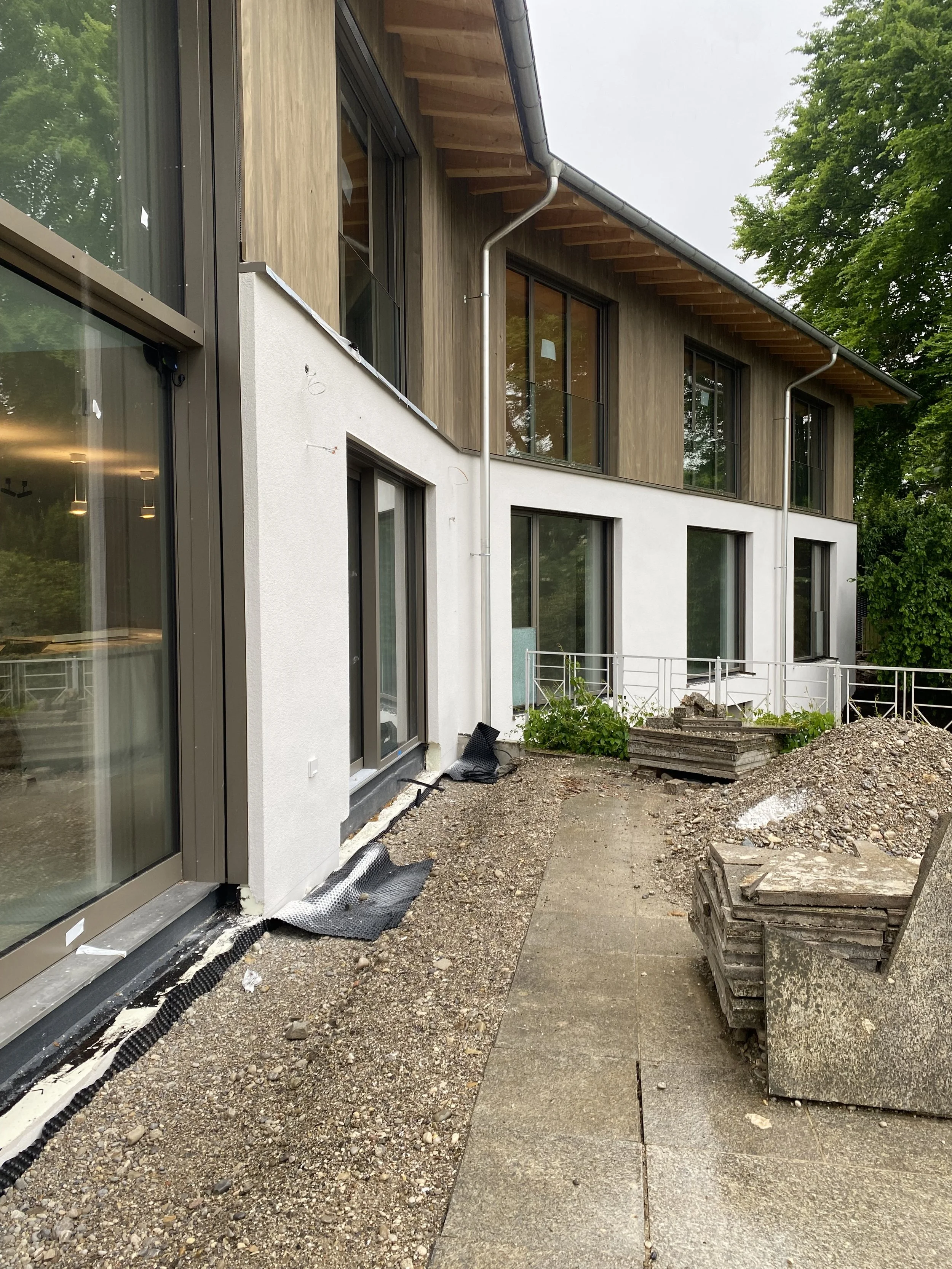 Bauarbeiten an einem modernen zweigeschossigen Gebäude mit großen Fenstern und Holzverkleidung, vor dem Gebäude befindet sich ein unbefestigter Weg, Baustellenmaterialien und Erde sichtbar.